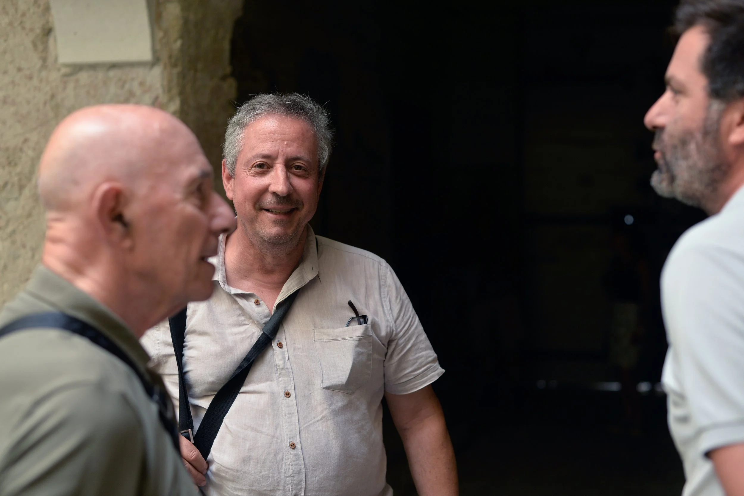 Trois hommes conversent dans un environnement sombre, éclairés en partie, avec un mur en pierre en arrière-plan.
