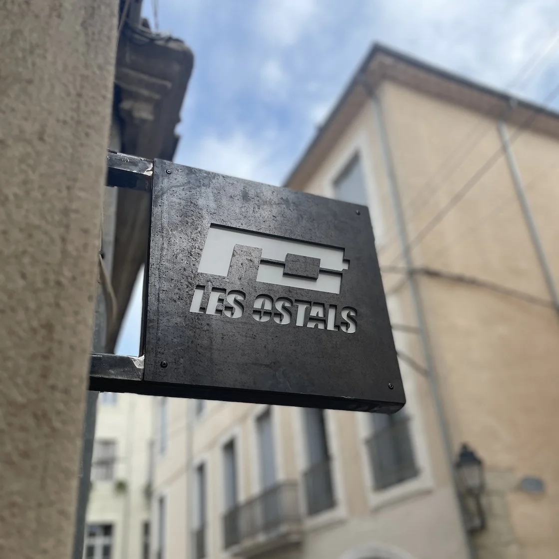panneau en métal noir avec logo stylisé et texte 'Les Ostals' fixé à un mur extérieur, avec bâtiments et ciel en arrière-plan