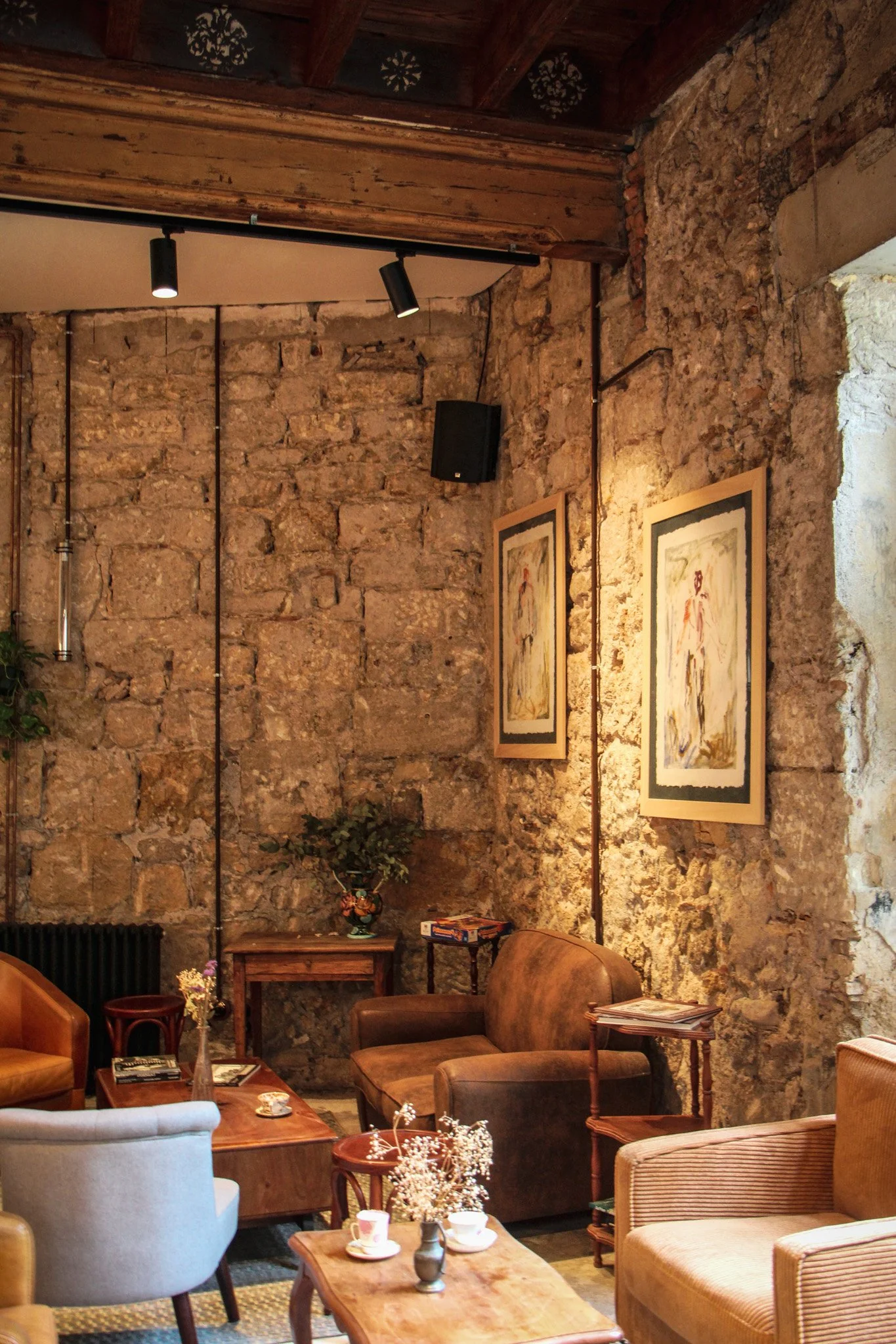 Salon avec murs en pierre, fauteuils en cuir, peintures encadrées sur le mur, tables en bois, décor vintage et ambiance chaleureuse.
