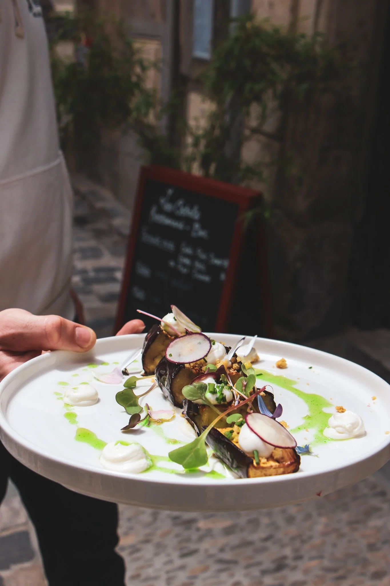 Une assiette de légumes grillés décorée de microgreens, de radis et de sauces végétales, tenue par une personne devant un fond de restaurant ou café avec des panneaux et des plantes en arrière-plan.