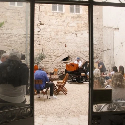 Groupe de personnes assises dans une cour extérieure, regardant un musicien qui joue de la musique près d'une vieille pierre et d'une armoire orange.
