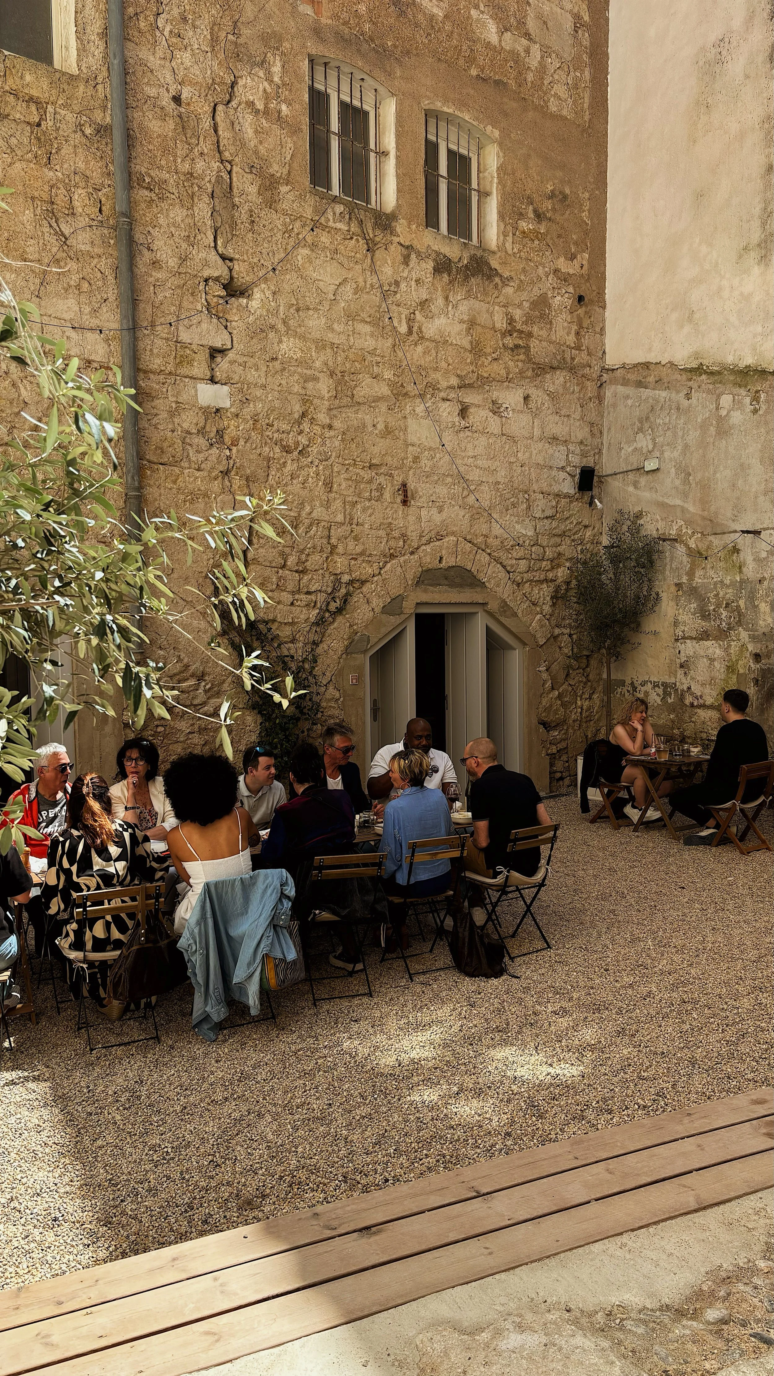 Groupe de personnes assises à une table de restaurant en plein air, devant un mur en pierre ancienne avec deux fenêtres.