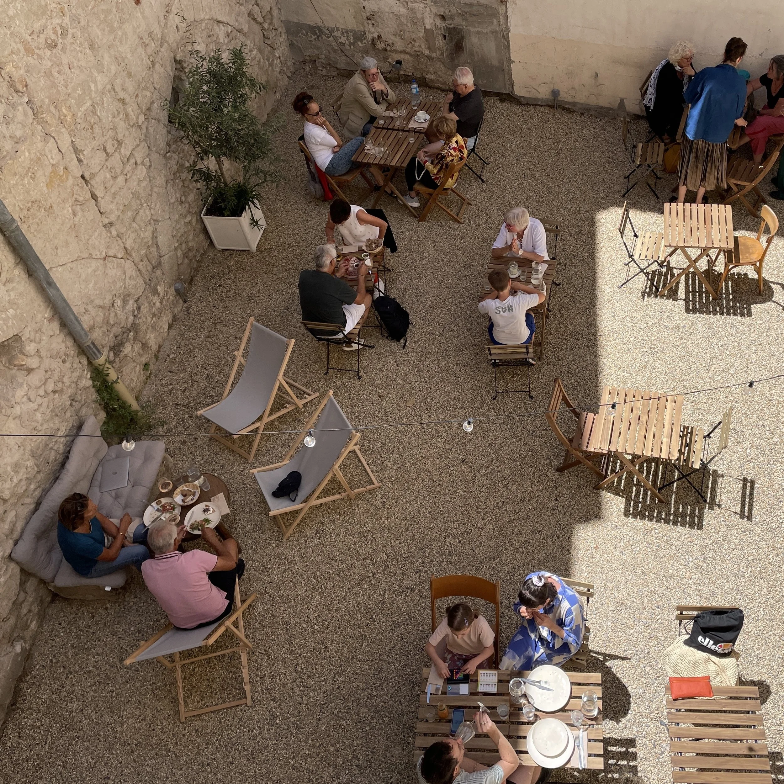Des personnes assises à des tables en terrasse, en train de manger et de discuter. L'espace est en plein air, entouré de murs en pierre et de mobilier varié.