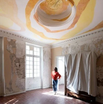 Une personne regarde par une fenêtre dans une pièce en rénovation, avec des murs partiellement dénudés et un plafond peint avec des tons jaunes et orangés.