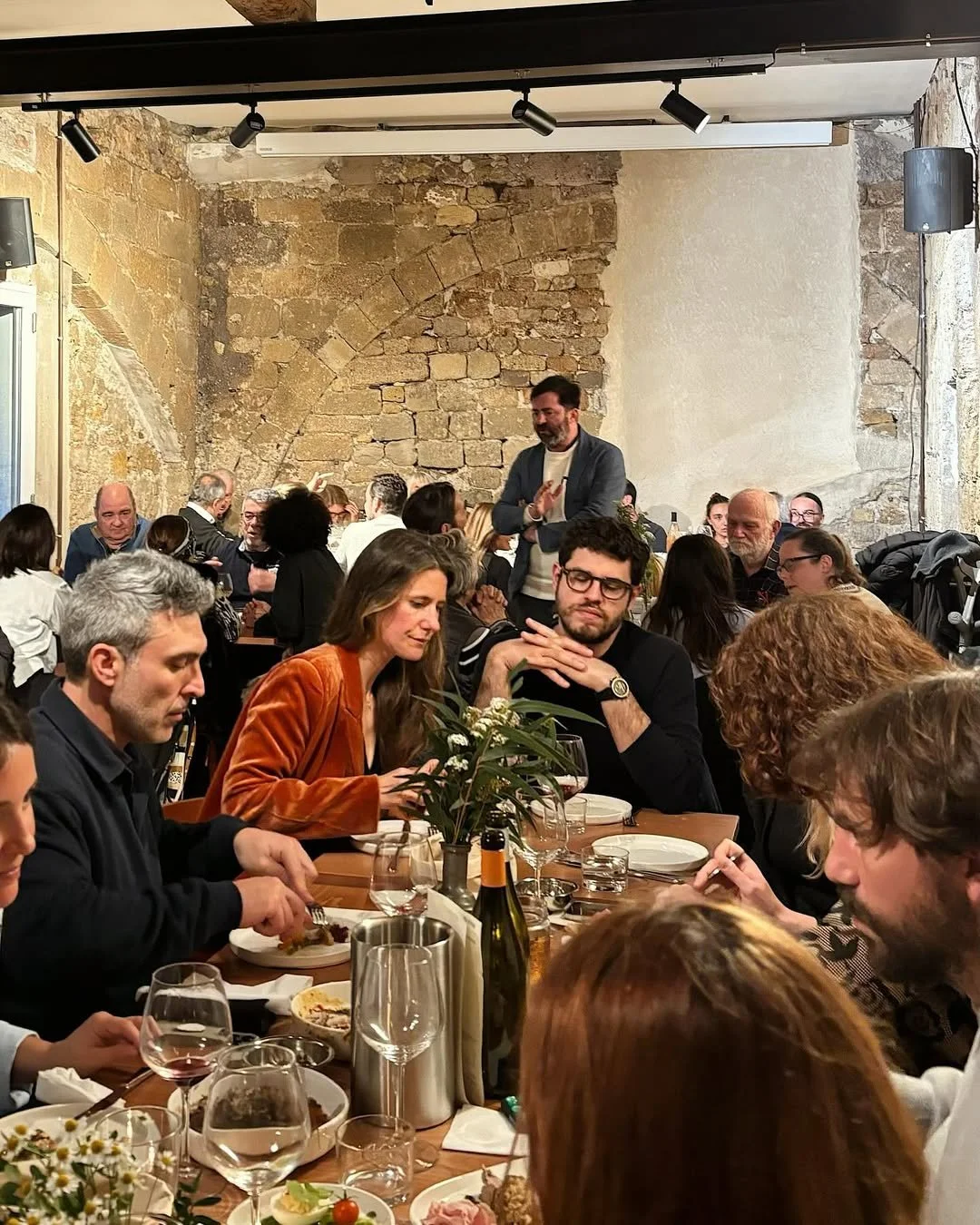 Groupe de personnes assises autour d'une table à un repas dans un restaurant aux murs en pierre, ambiance conviviale et chaleureuse.