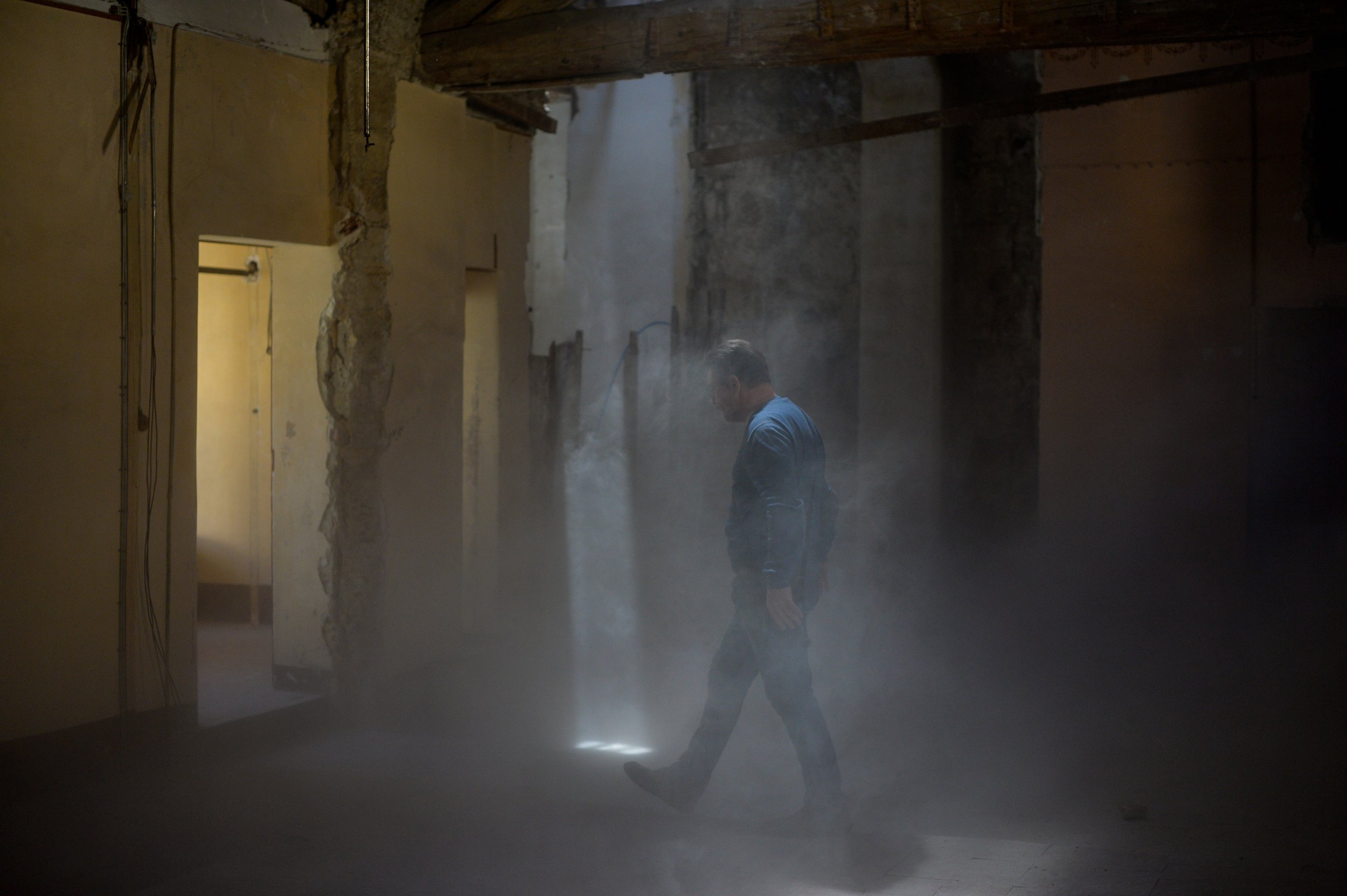 Un homme marche dans une pièce sombre remplie de poussière ou de fumée, avec des murs partiellement démolis dans un bâtiment en rénovation.