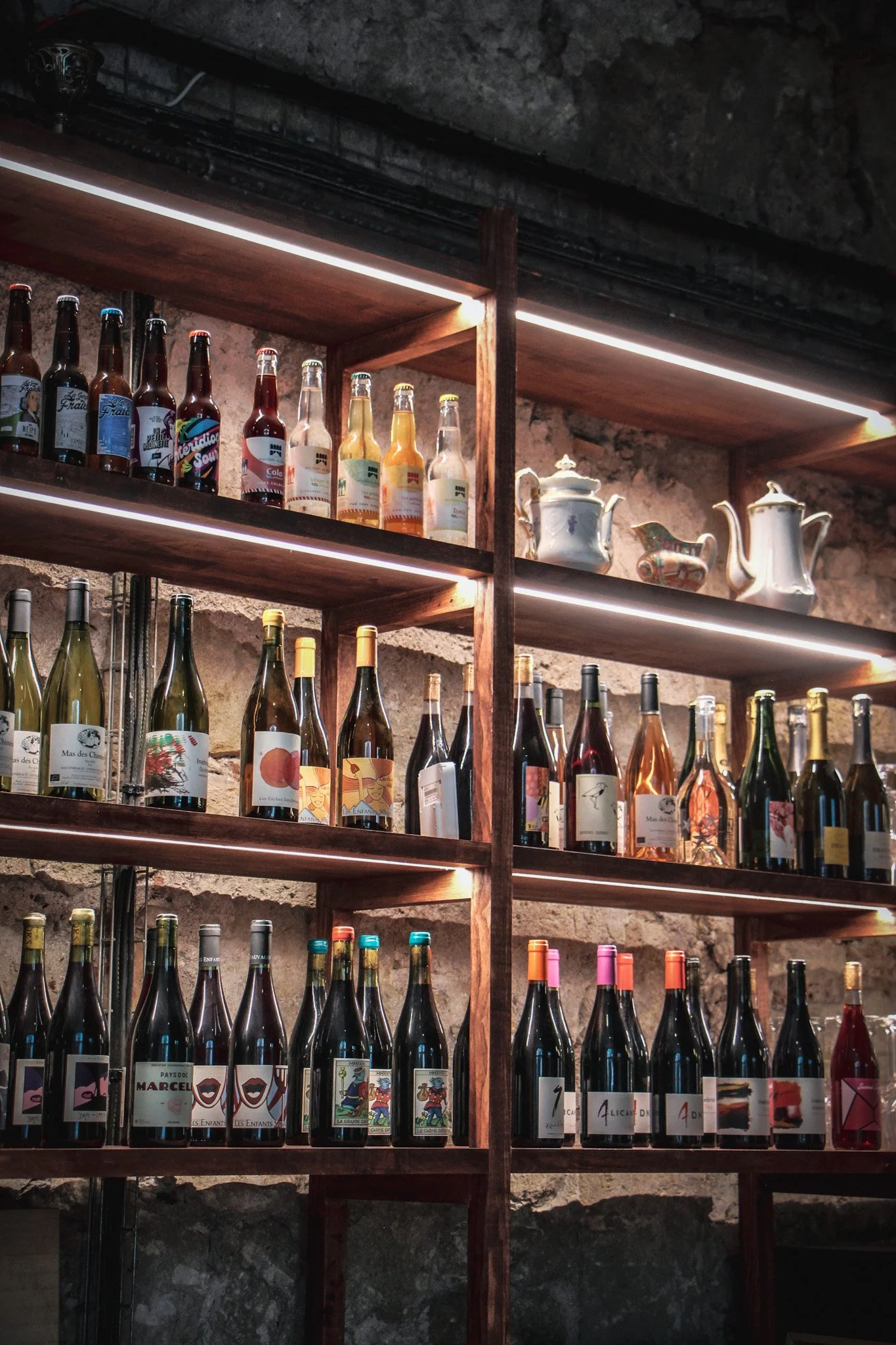 Étagère en bois avec bouteilles de vin et boissons non alcoolisées, ainsi que des théières en porcelaine, contre un mur en pierre, éclairée par des lumières LED intégrées.