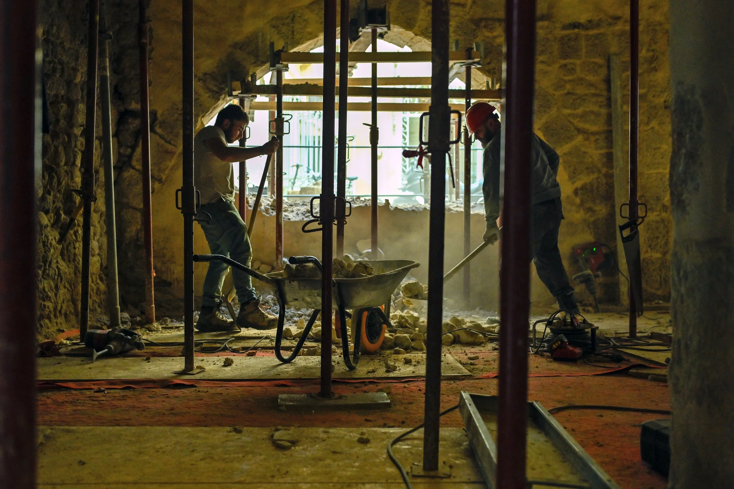 Deux ouvriers construisent ou rénovent une structure en pierre à l'intérieur d'une pièce, utilisant des outils tels que pelles et marteaux, avec des escabeaux et un support en acier. Il y a de la poussière, et un grand trou dans la muraille laisse pa