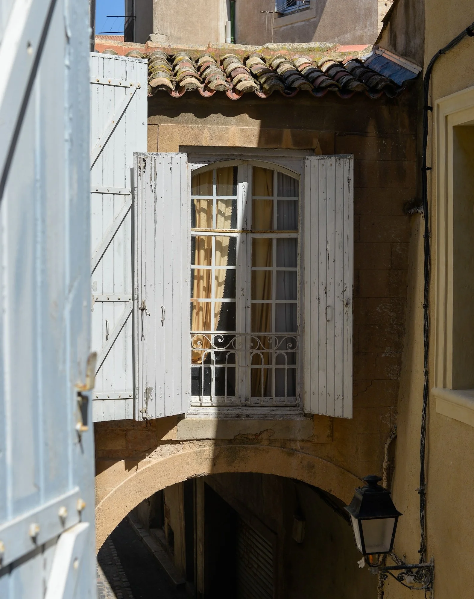 Fenêtre avec volets en bois blanc, rideaux beige derrière la vitre, extérieur en pierre et tuiles au toit, lampadaire en fer noir sur le côté, ruelle sombre en bas sous une arche. 