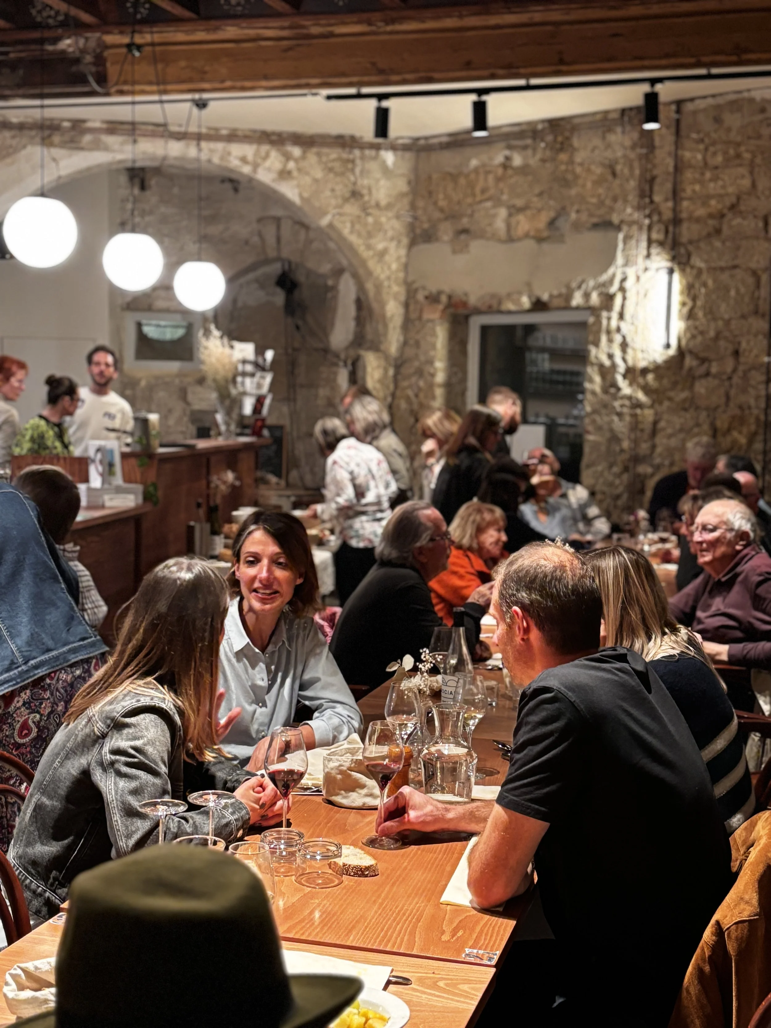 Groupe de personnes assises autour d'une table en intérieur, discutant et dégustant du vin, dans un cadre rustique avec murs en pierre et éclairage chaleureux.