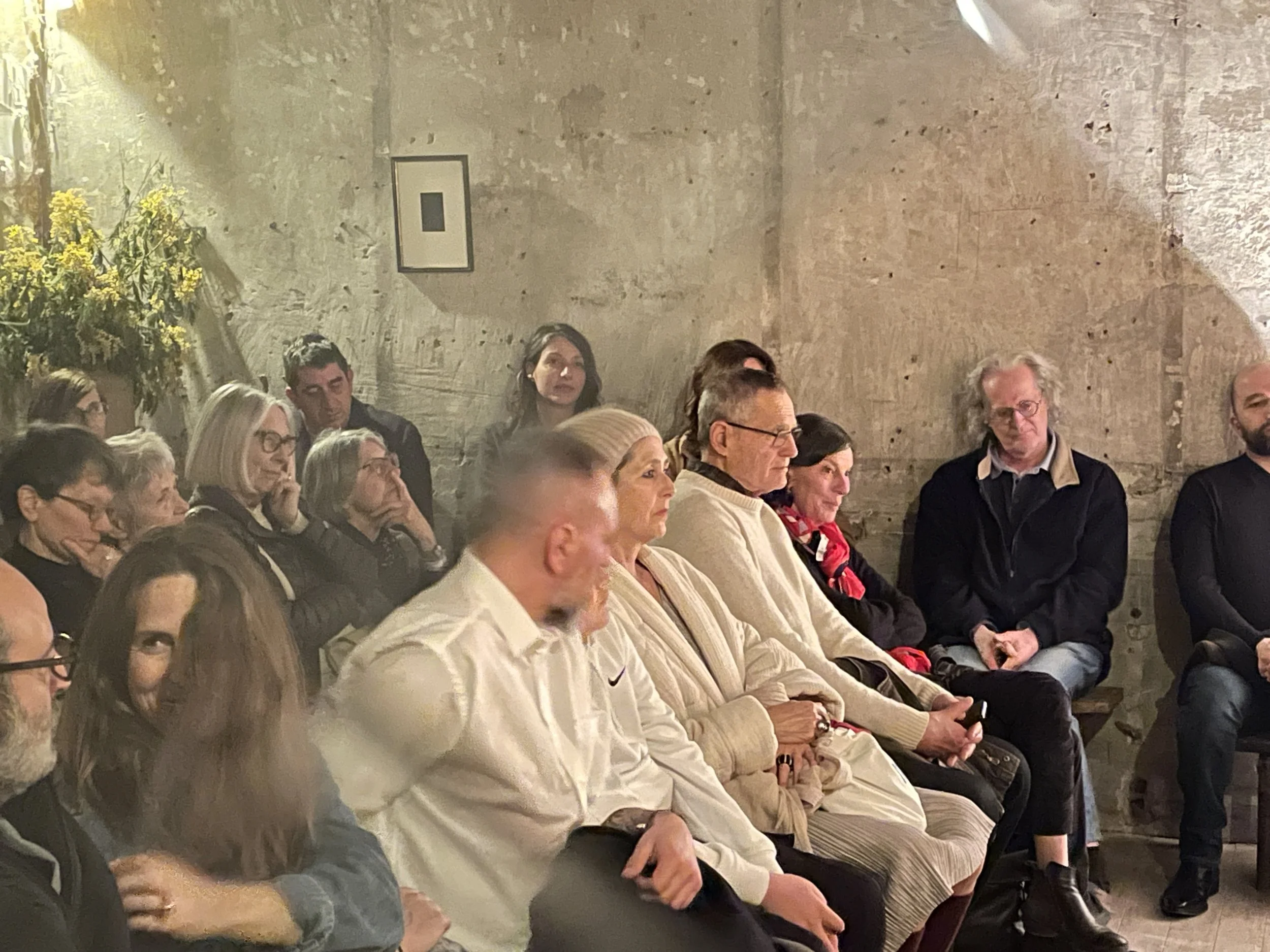 Un groupe de personnes assises en rangée dans une pièce aux murs en pierre, écoutant attentivement un discours ou une présentation.