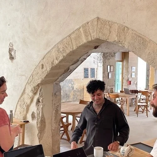 Trois personnes souriantes dans un café, avec une arche en pierre derrière elles, et des tables en bois avec des chaises.