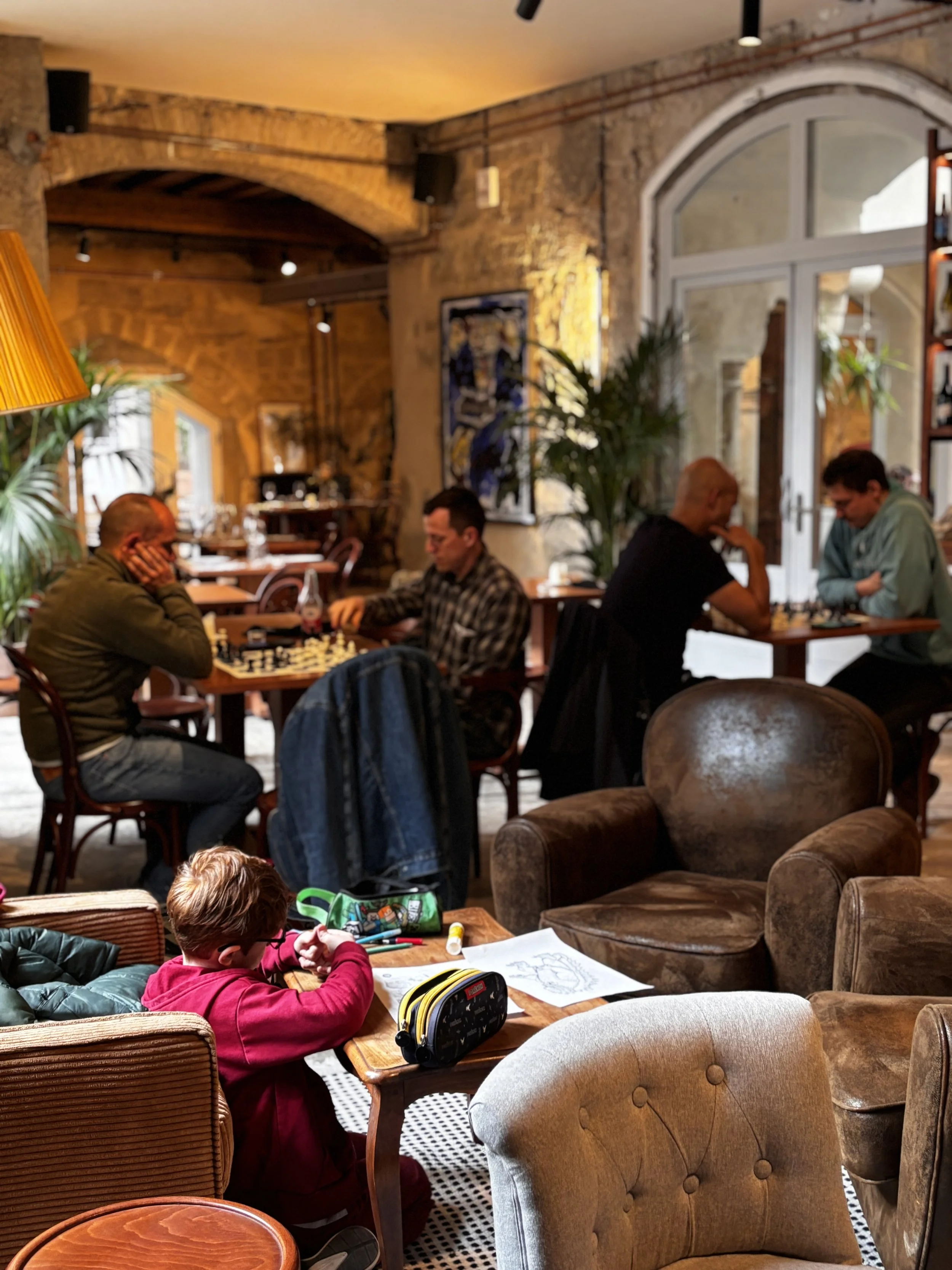 Une scène intérieure d'un café ou lounge avec des personnes, dont un enfant qui dessine, et d'autres adultes jouant aux échecs, dans une ambiance chaleureuse et conviviale.