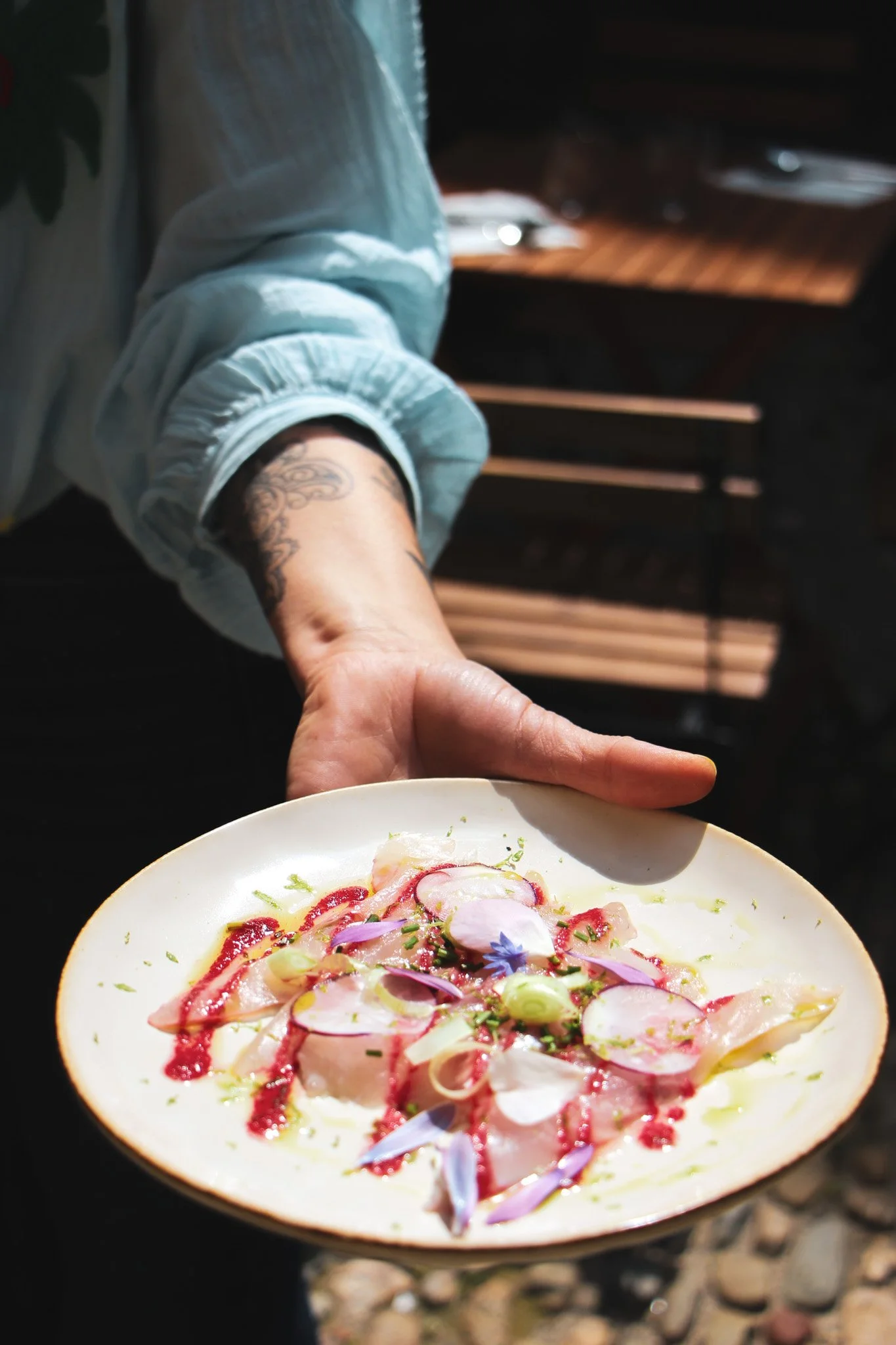 Assiette de carpaccio de poisson avec des garnitures végétales, tenu par une personne dont le bras tatoué, en arrière-plan un intérieur en bois.