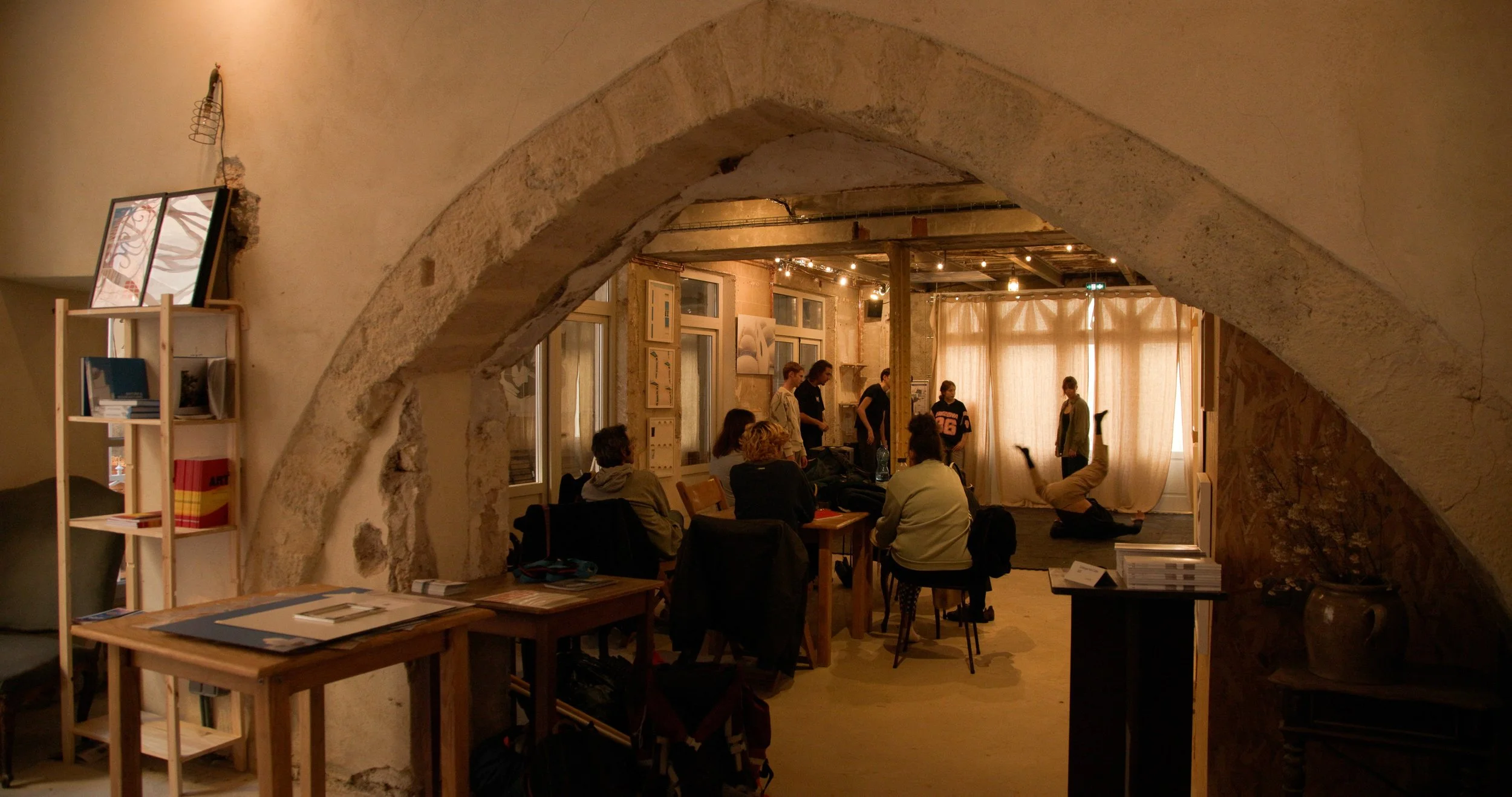 Intérieur d'un espace de coworking avec des personnes assises et discutant, un tableau au mur, et une personne faisant de la danse ou du yoga près de rideaux au fond.