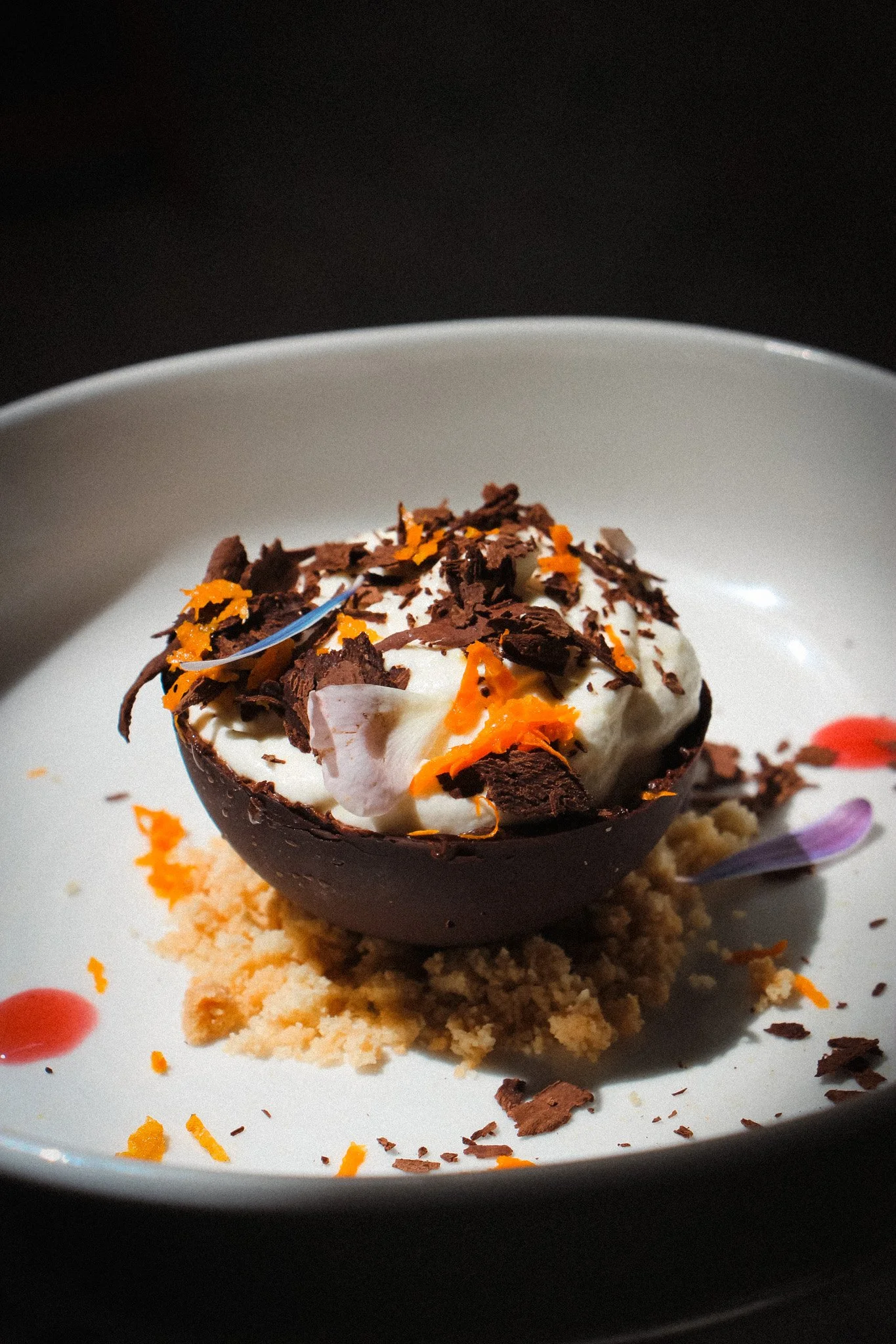 Dessert présenté dans une coupe en chocolat, avec une boule de glace, des copeaux de chocolat et des zestes d'orange, sur un lit de biscuit émietté, dans une assiette blanche.