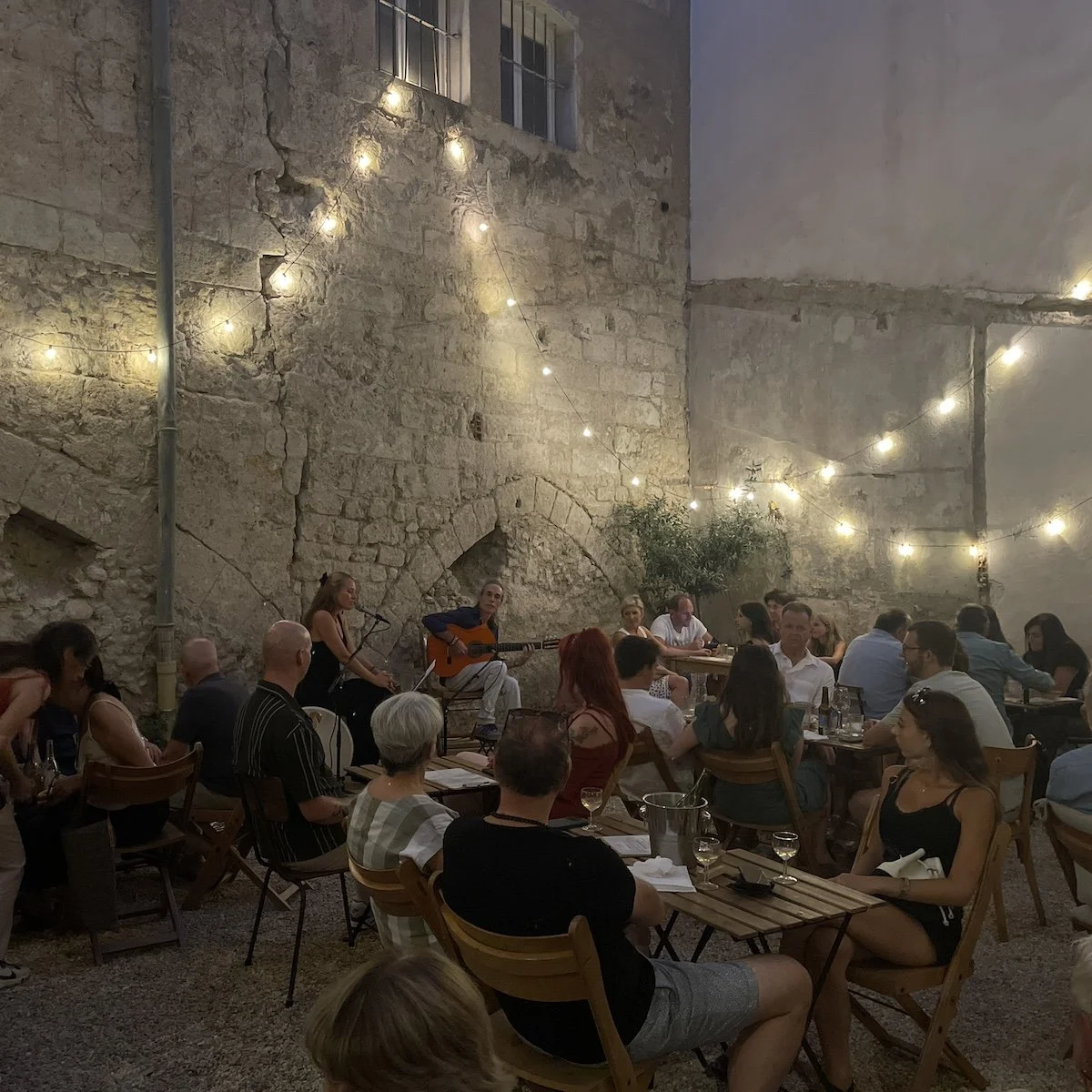 Groupe de personnes assises dans une cour intérieure avec un mur en pierre, éclairée par des guirlandes lumineuses, où deux musiciens jouent de la guitare.