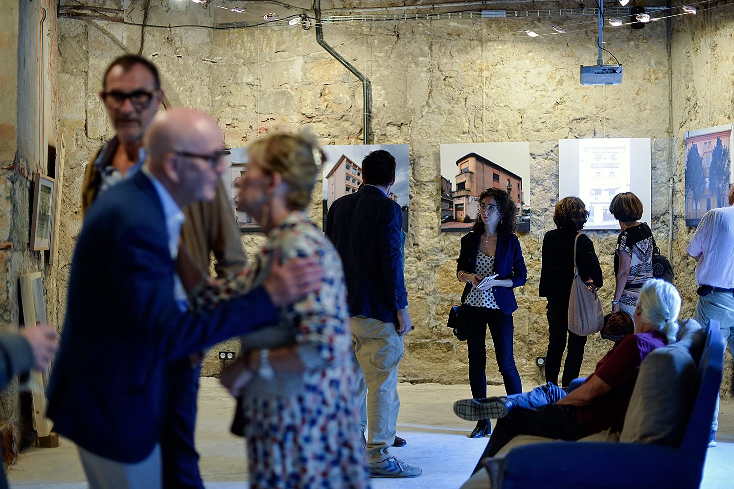Groupe de personnes lors d'une exposition d'art ou une visite guidée dans un espace avec des murs en pierre, regardant et discutant autour de photographies et d'œuvres d'art suspendues.