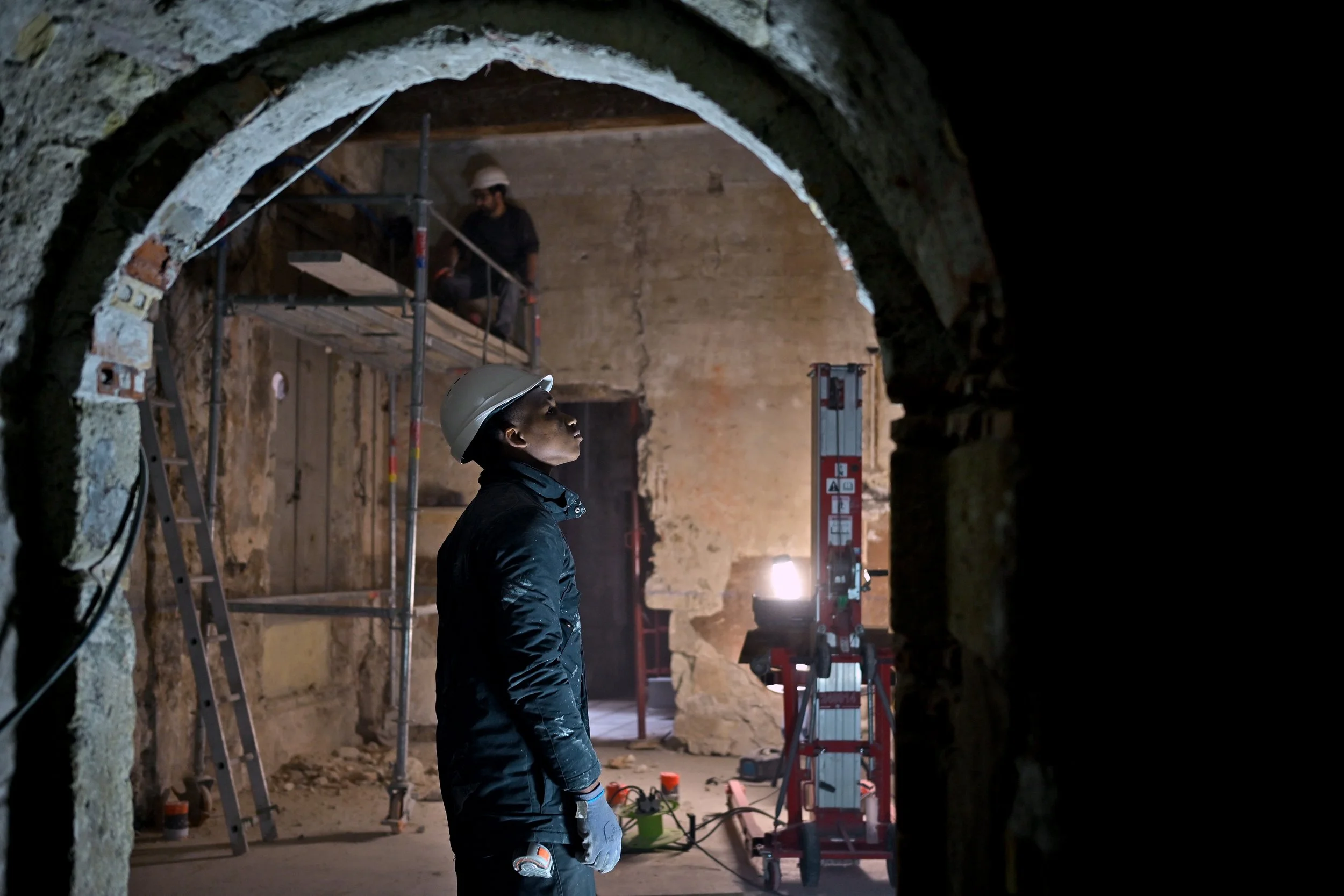 Un homme portant un casque de chantier regarde vers le haut dans une zone de construction souterraine, entouré d'échafaudages et de matériel de construction.