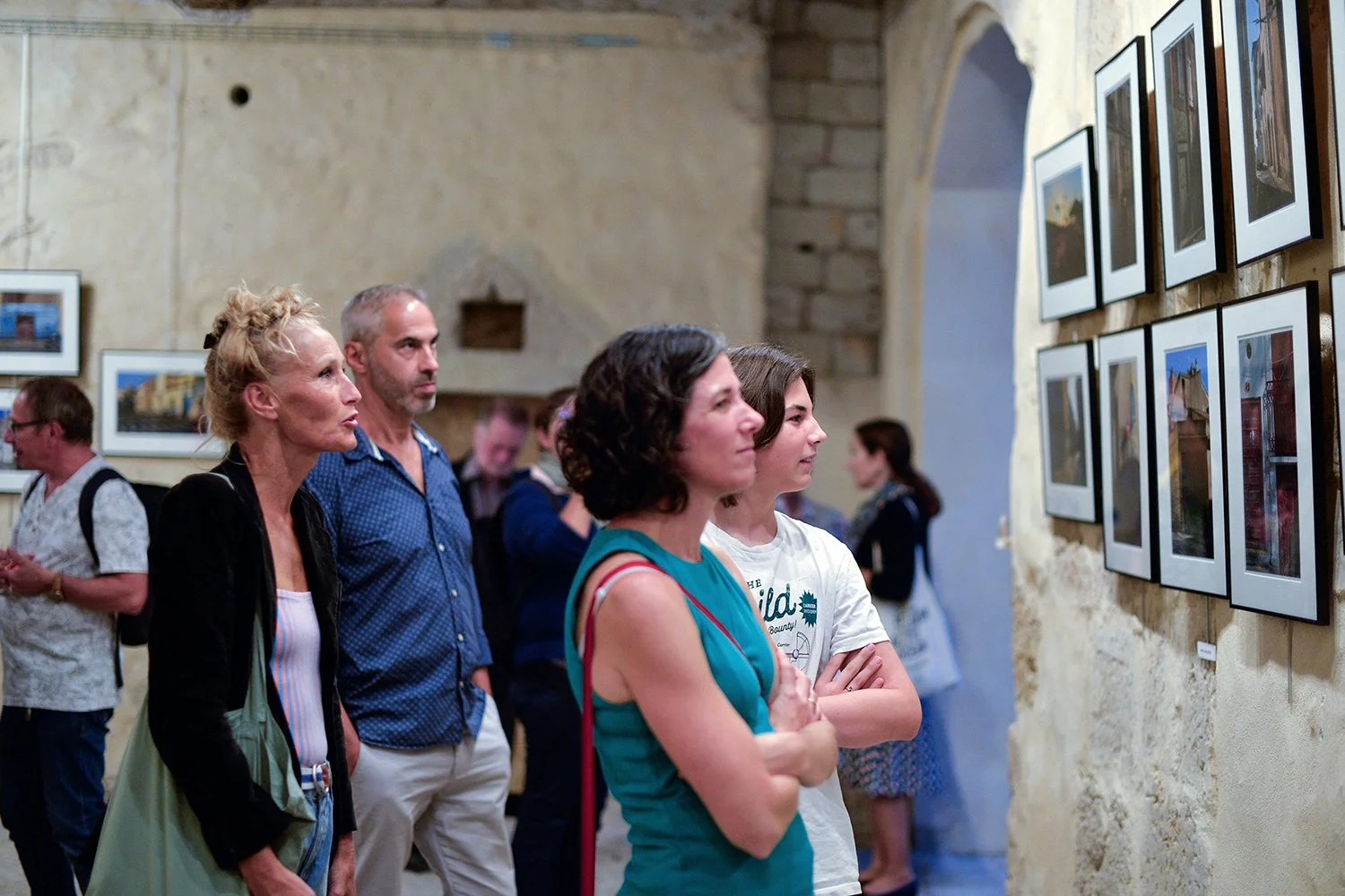 Groupe de personnes regardant des photographies dans une galerie d'art, murs en pierre et tableaux encadrés.