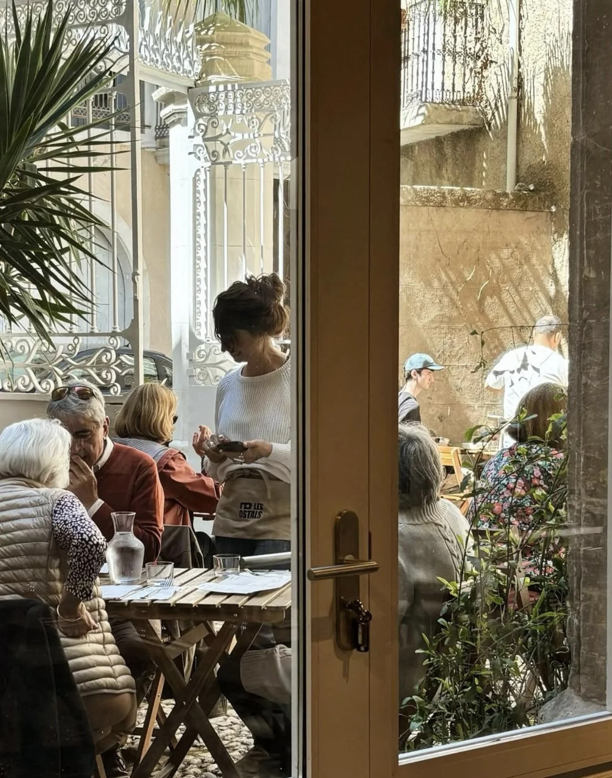 Groupe de personnes dans un café ou un restaurant, vue à travers une porte en verre, avec des décorations extérieures et des plantes.