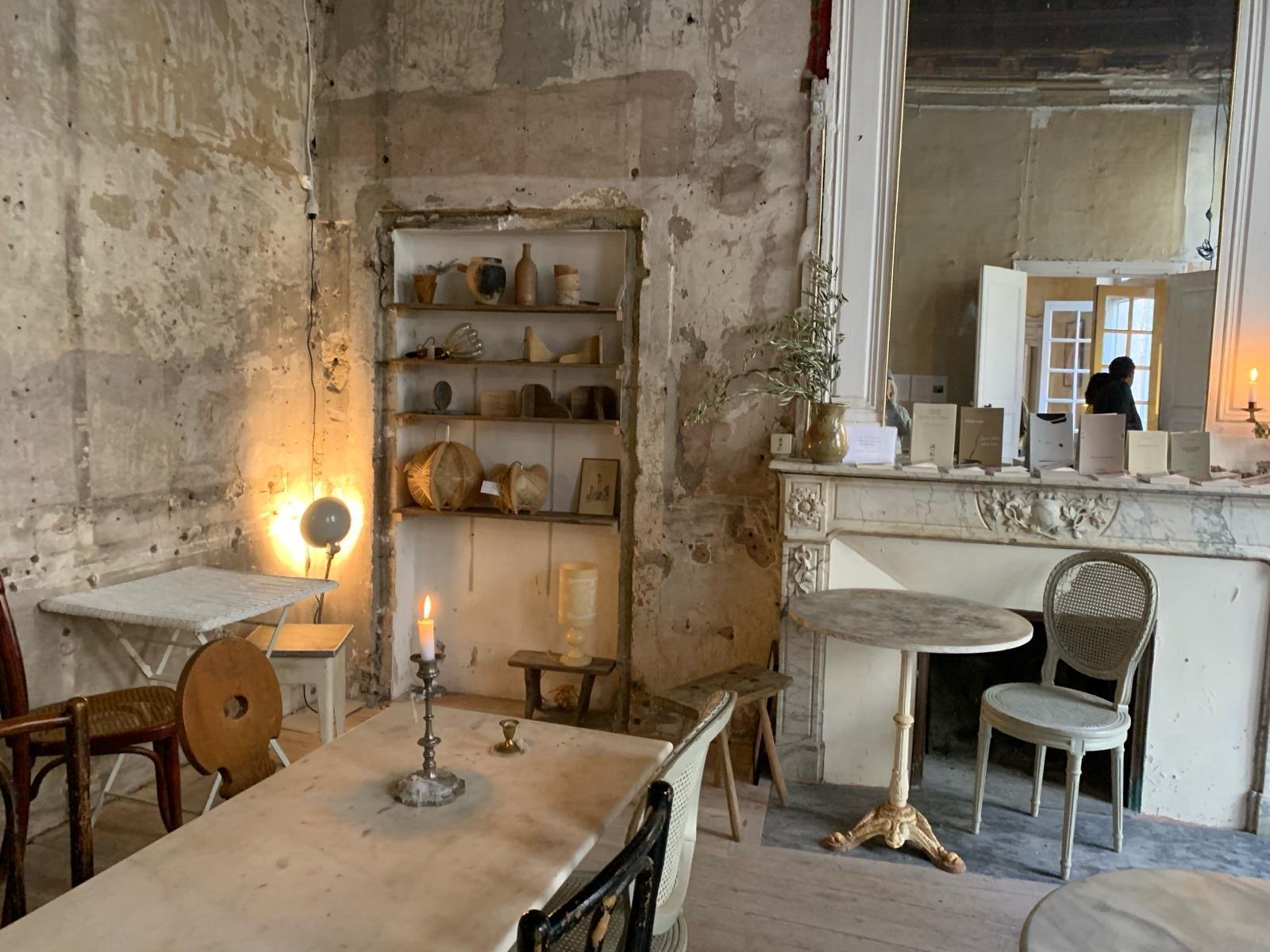 Intérieur d'un salon avec un mur en pierre, une cheminée en marbre, des tables et des chaises anciennes, une étagère en bois avec des objets en céramique, une bougie et une lampe sur une table.