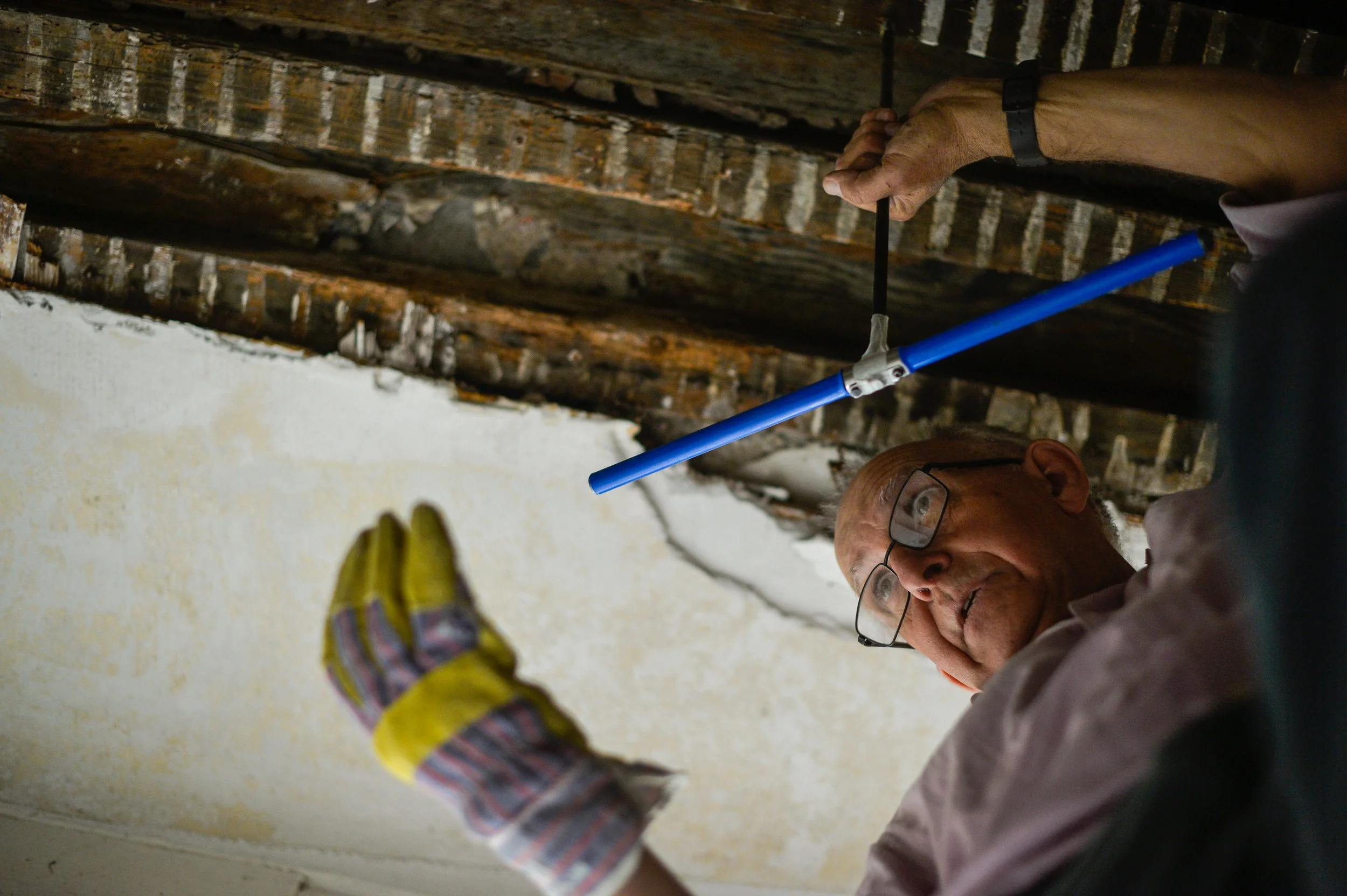 Un homme portant des lunettes et des gants de travail examine ou travaille sur un plafond en bois, utilisant un outil bleu en forme de perche ou de tige.