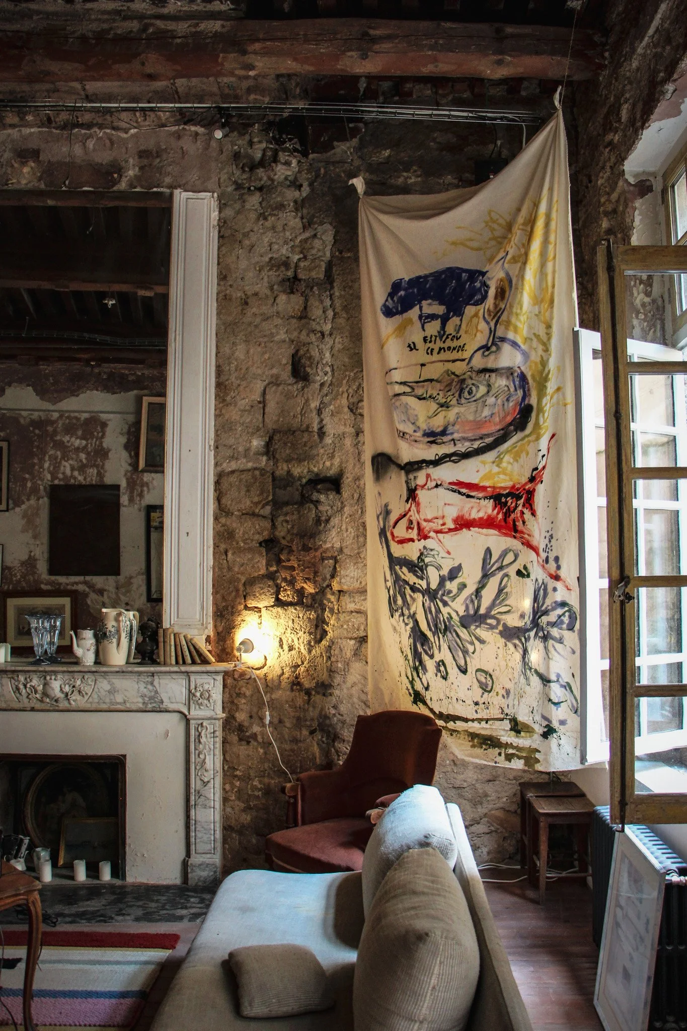 Intérieur d'une pièce avec un mur en pierre, une cheminée ancienne, un fauteuil marron, un canapé beige, une lampe, une grande fresque ou panneau suspendu avec des dessins colorés, et une fenêtre avec des volets ouverts.