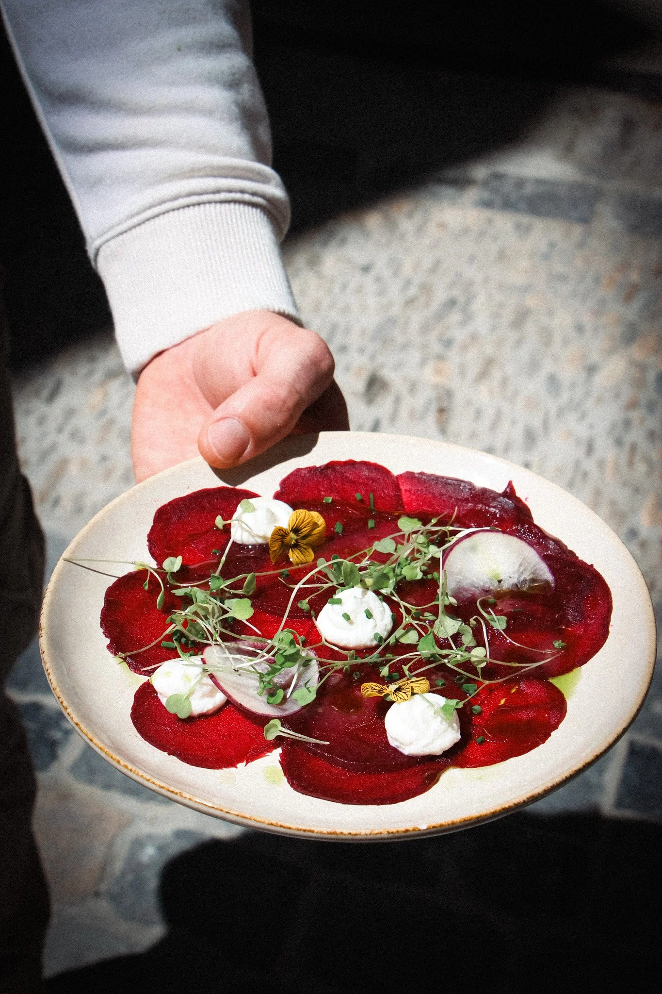 Une personne tient une assiette de tartare de betteraves décoré de microgreens, de petites meringues et de fleurs comestibles, sur fond sombre.