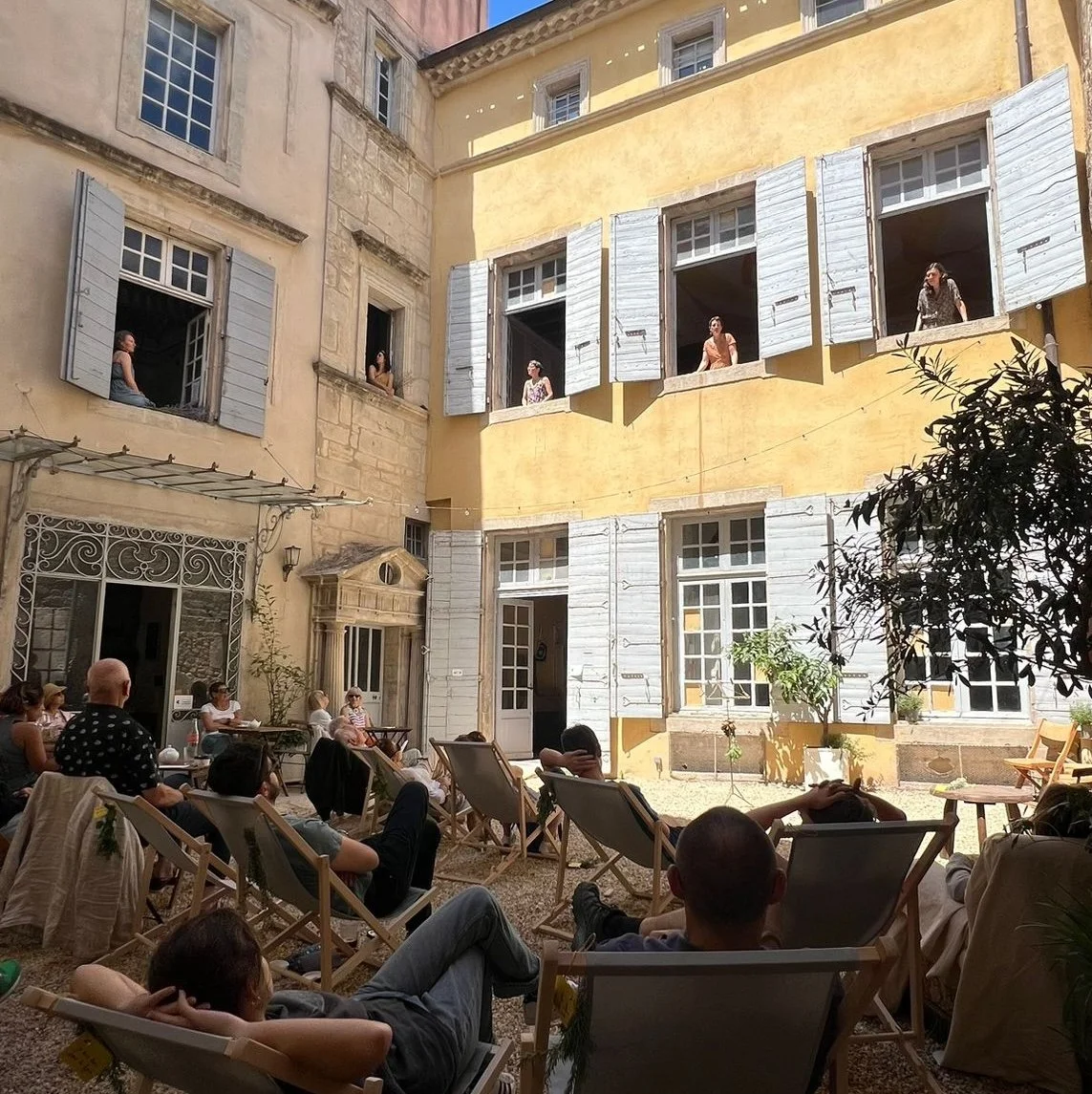 Un groupe de personnes assises et se relaxant dans une cour intérieure, regardant un film ou une présentation sur un écran, entouré de bâtiments anciens avec fenêtres à volets bleus ouverts.