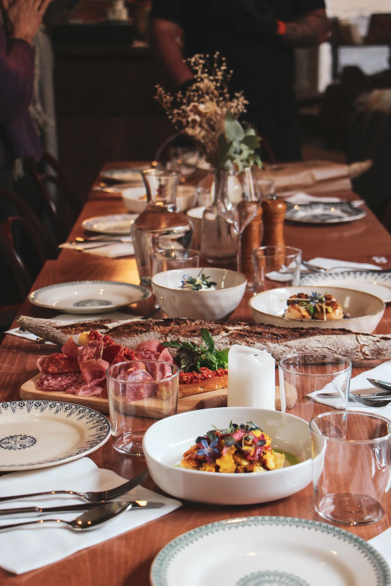 Table de restaurant élégamment dressée avec des plats de charcuterie, une pizza, un plat de pâtes, des verres d'eau, une bougie et un vase avec des fleurs séchées.