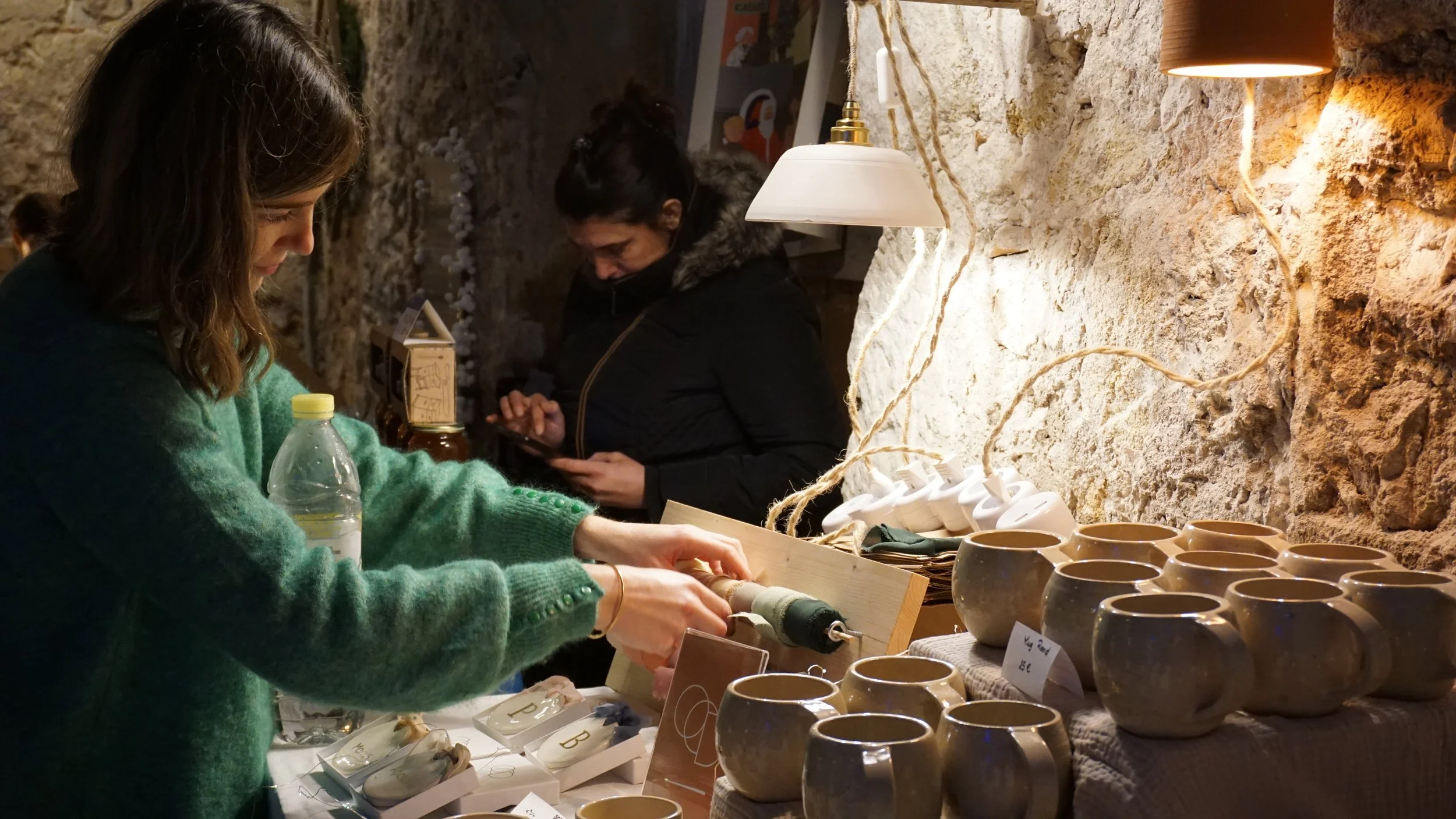 Deux femmes vendant des objets en céramique dans un marché artisanal, avec des tasses et des décorations en terre cuite sur un stand en pierre.
