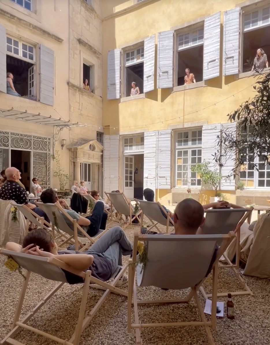 Une scène extérieure dans un patio avec des personnes assises sur des chaises longues, regardant un spectacle ou un film projeté sur un mur de bâtiment jaune avec des volets en bois blanc. Des femmes regardent depuis des fenêtres ouvertes.