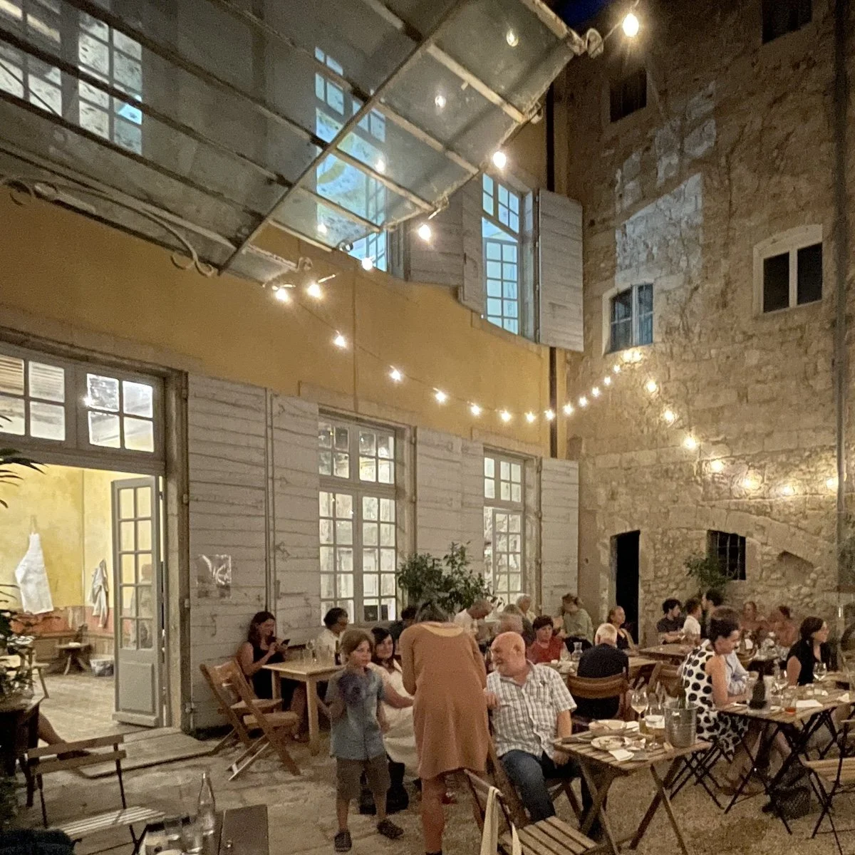 Un groupe de personnes mangeant et socialisant dans un restaurant ou une salle à manger avec des murs en pierre et des fenêtres à plusieurs volets, décoré avec des guirlandes lumineuses.