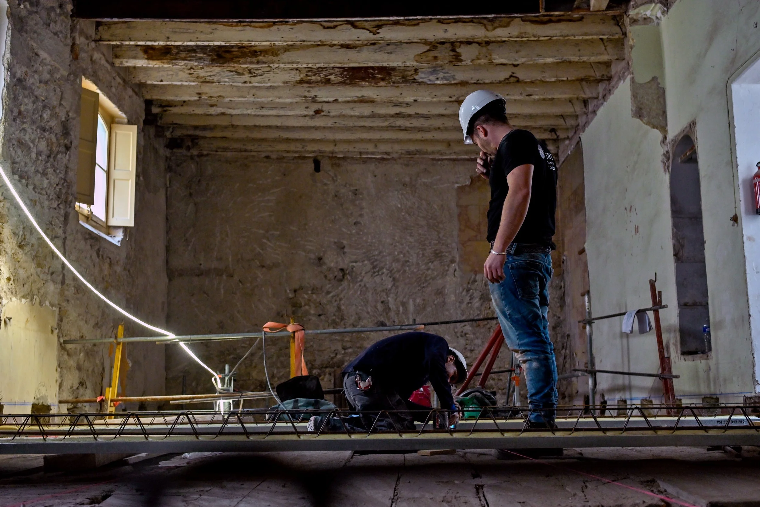 Deux travailleurs dans une pièce en rénovation, un portant un casque de sécurité et observant l'autre qui travaille au sol, avec des éléments de construction autour.