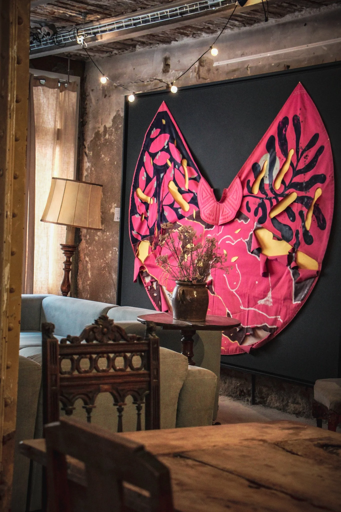 Un salon avec un tableau mural représentant un papillon stylisé en tissu rose, noir et jaune. Décor rustique avec meubles en bois, un lampadaire avec abat-jour beige, et une table ronde avec un vase de fleurs séchées.