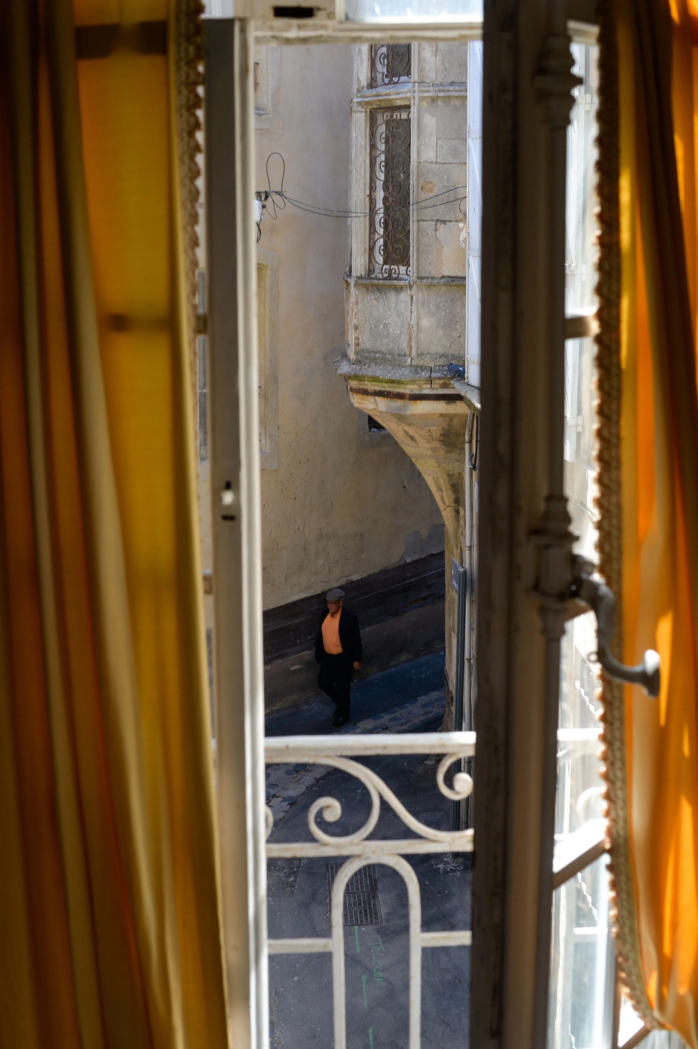 Photo vue de la fenêtre ouverte avec des rideaux jaunes, montrant une rue pavée avec un homme marchant.