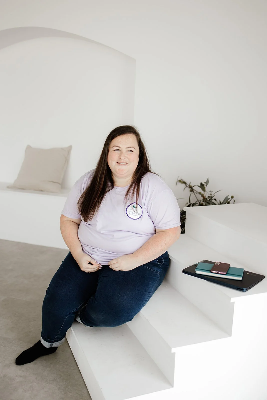 Megan, founder of The Wee Thistle Sleep Consultant, seated on white steps with a laptop and notebooks, wearing her branded lavender t-shirt.
