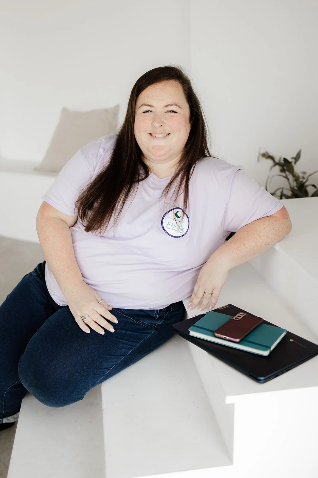 A woman with long dark hair, smiling, wearing a white T-shirt with a circular logo, sitting on a white table with a laptop, notebooks, and smartphone nearby, in a bright room with a white wall and some plants in the background.
