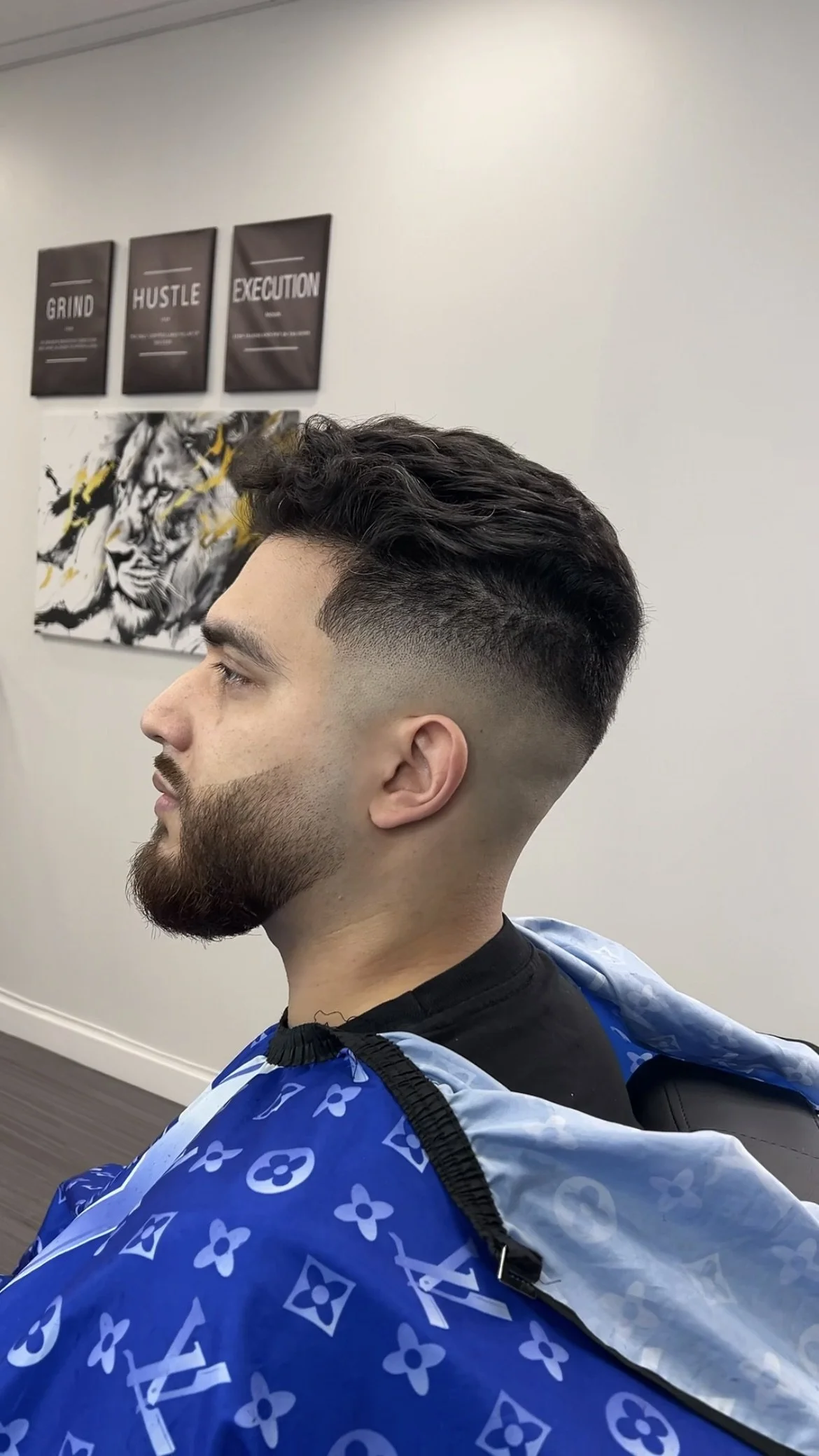 Un homme assis dans un salon de coiffure avec une coupe de cheveux dégradée, portant une cape bleue avec des motifs blancs. Au mur, il y a des affiches motivantes et un tableau artistique.