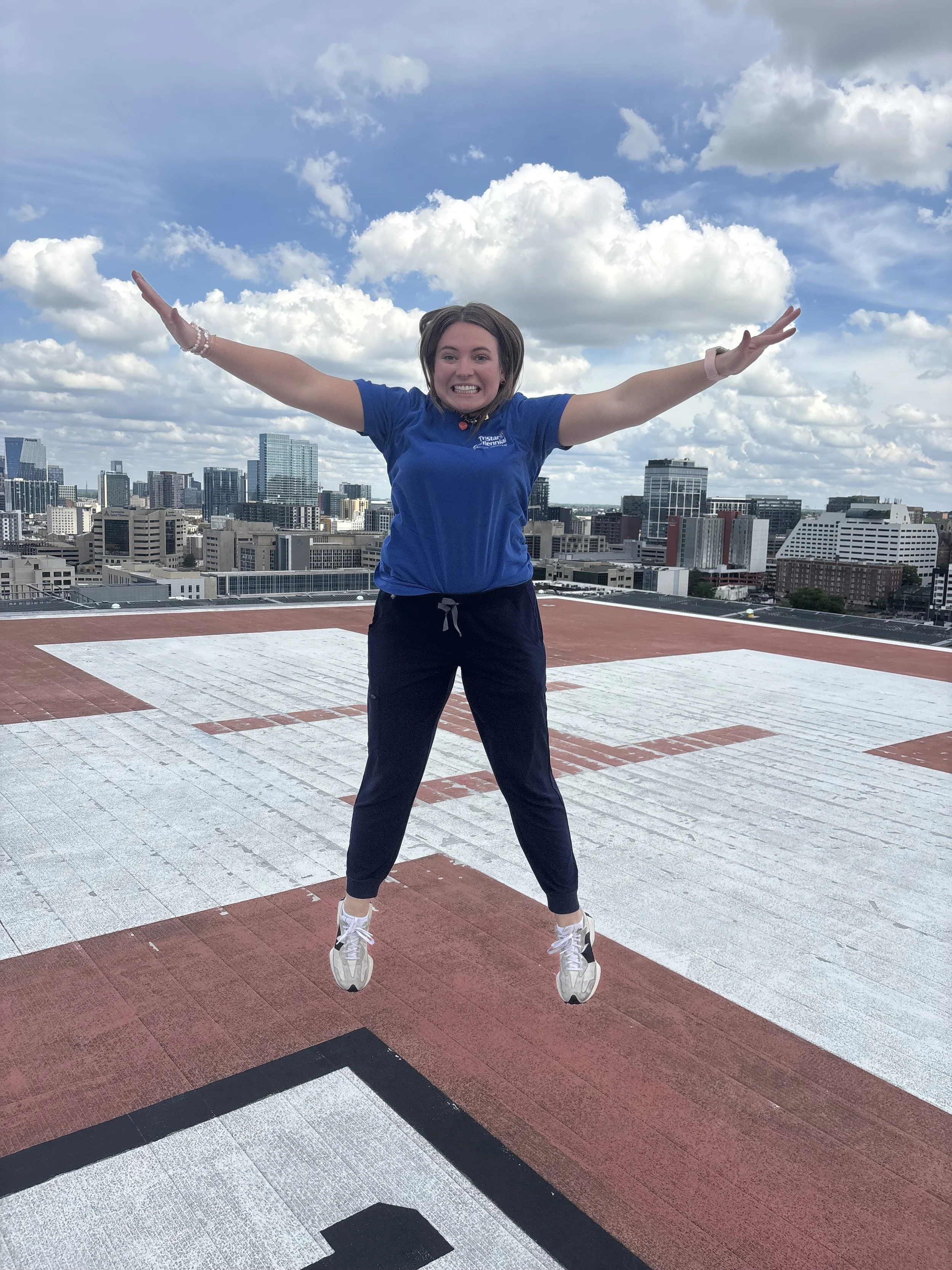 Young woman jumping with arms outstretched on a rooftop with city skyline and blue sky with clouds in the background.