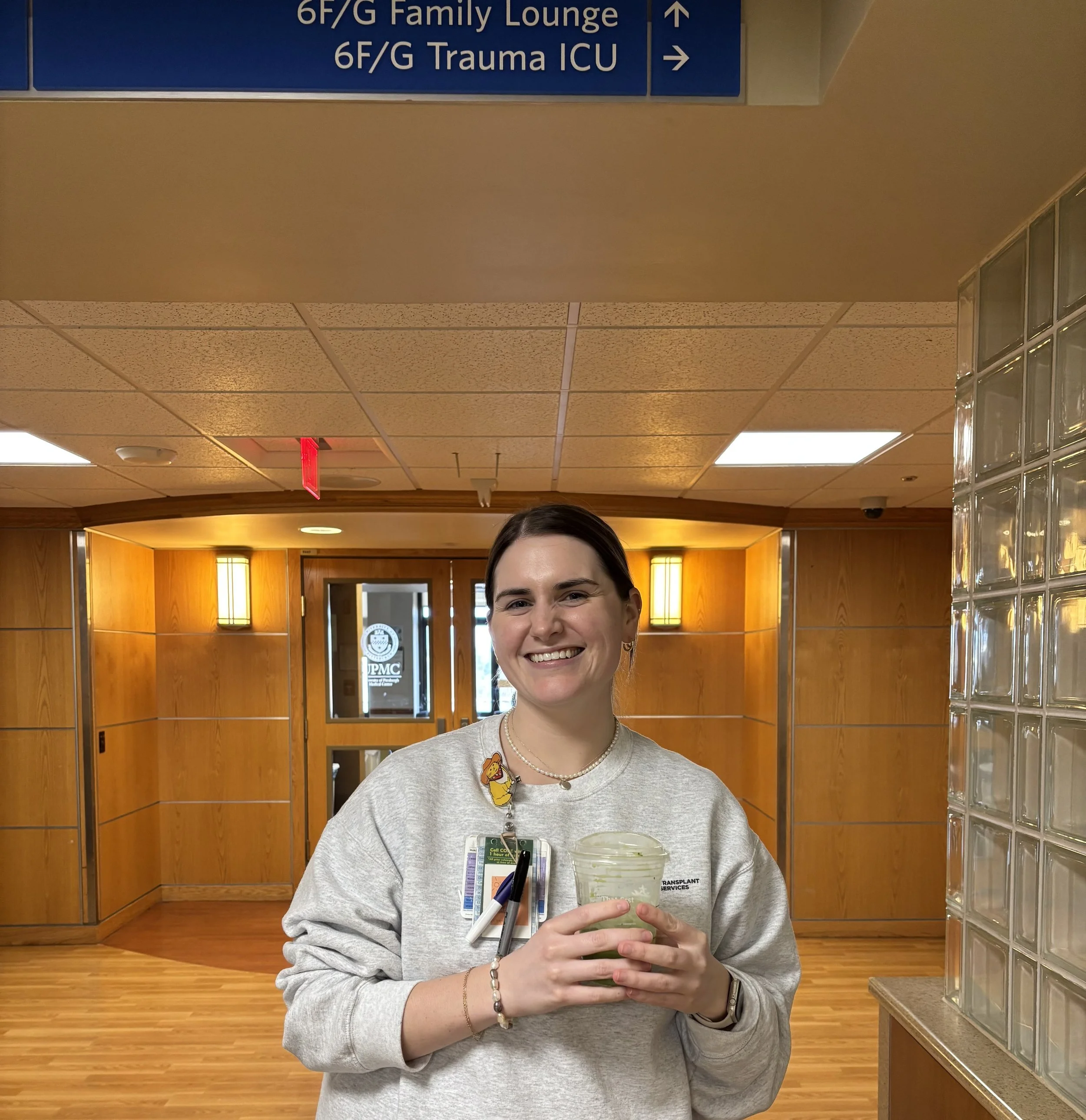 A young woman with dark hair smiling, holding a container of food, standing in a hospital corridor with wooden paneling and a sign overhead indicating directions to the Family Lounge and Trauma ICU.