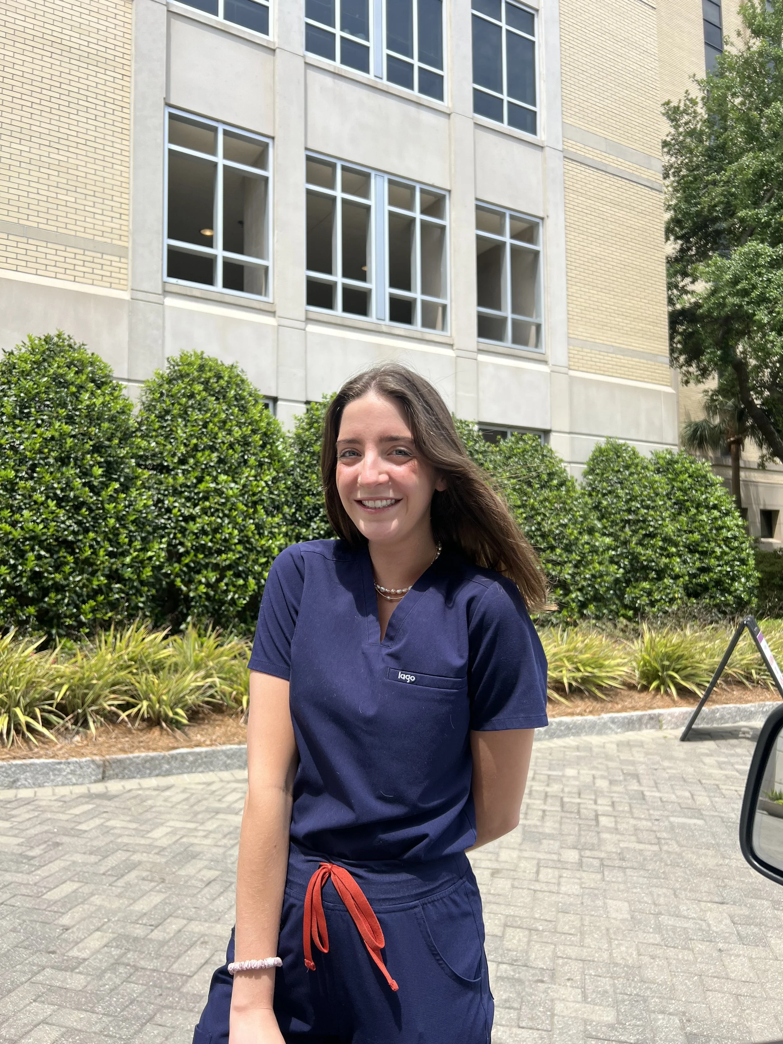 A woman in navy medical scrubs standing outdoors in front of green bushes and a modern building with large windows, smiling at the camera.