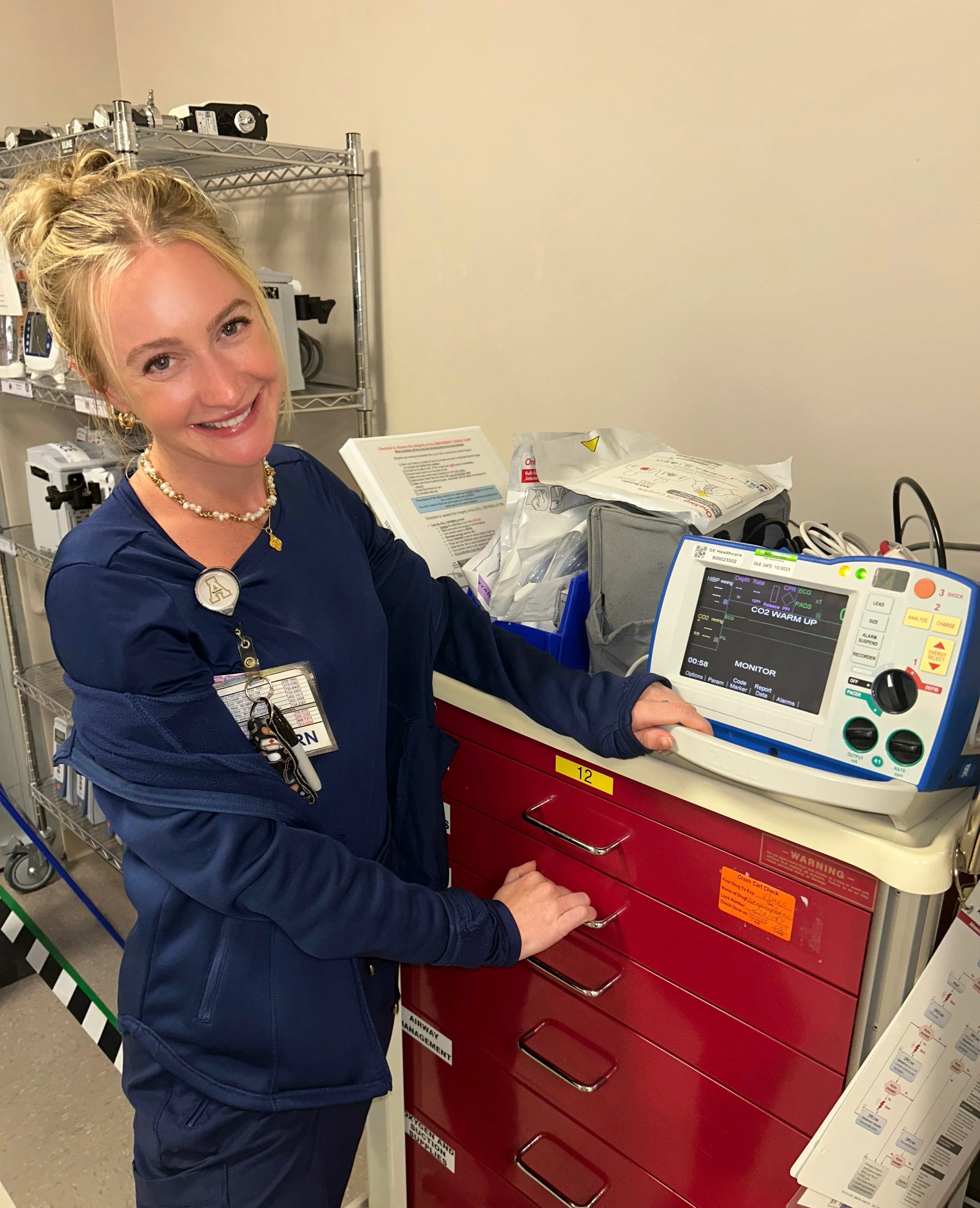 A smiling female nurse standing next to a medical monitor in a hospital setting.