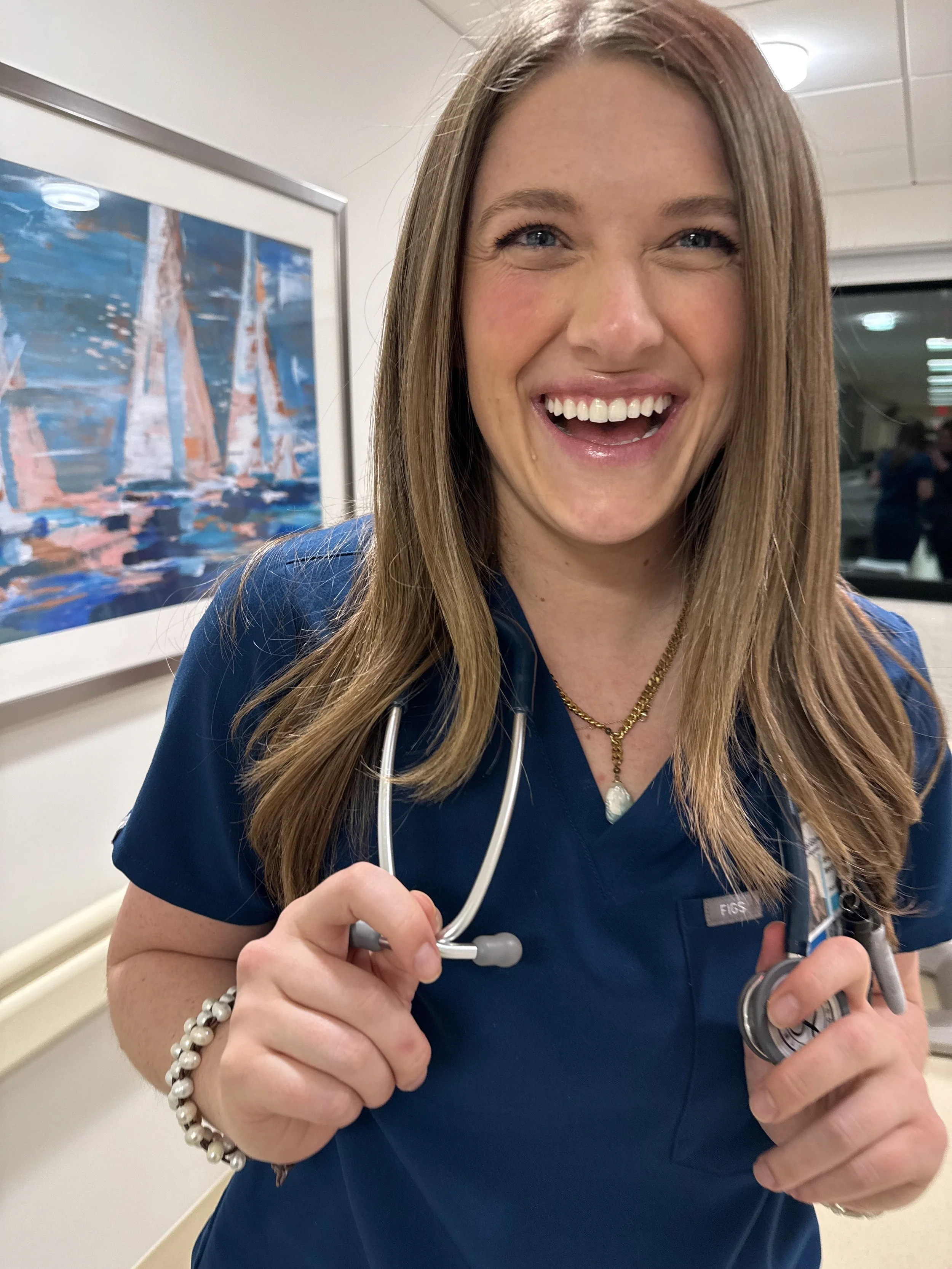 A smiling female healthcare professional wearing navy scrubs, holding a stethoscope, in a medical setting.