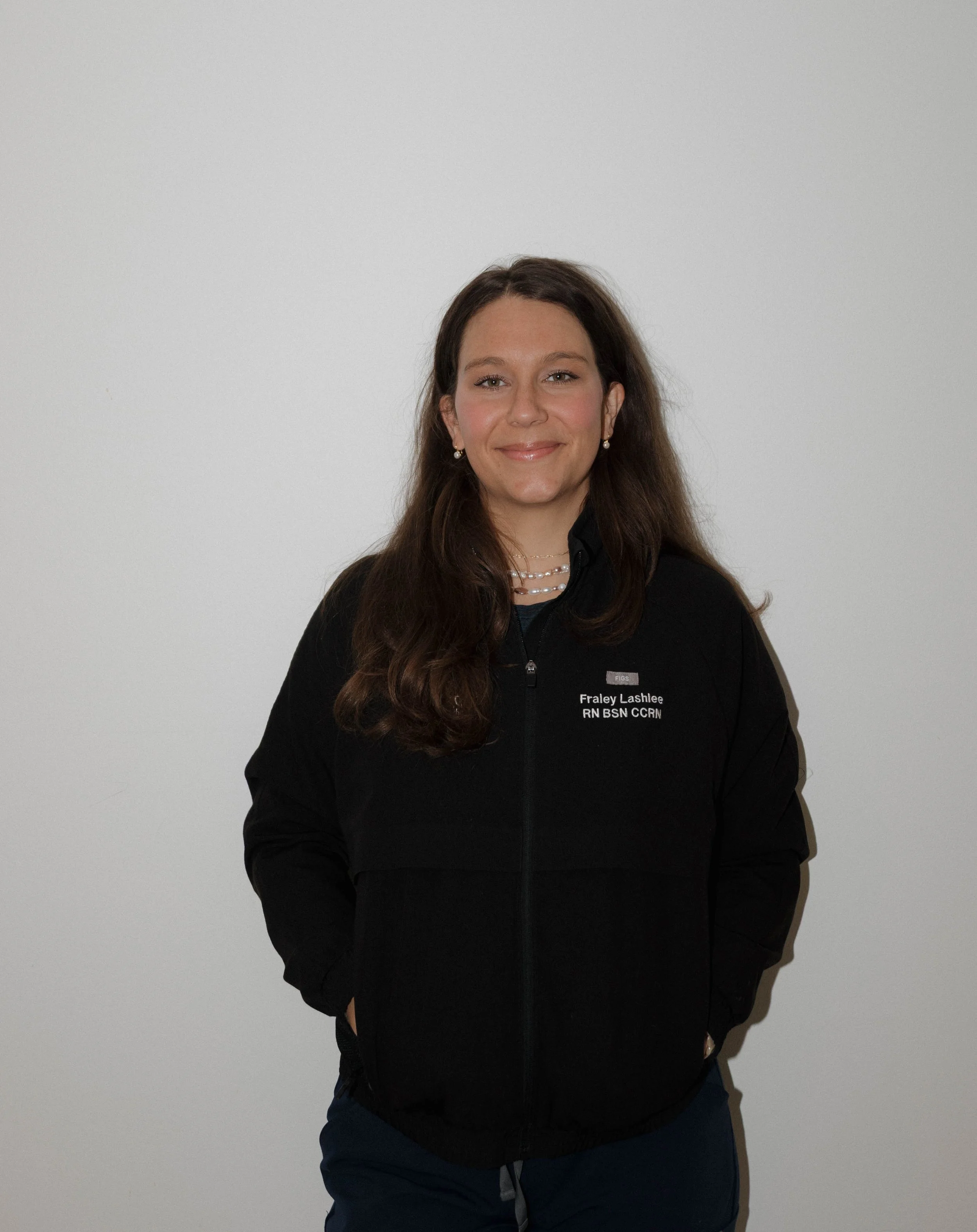 A woman with long brown hair smiling, wearing a black zip-up jacket with name tag, standing against a plain white wall.