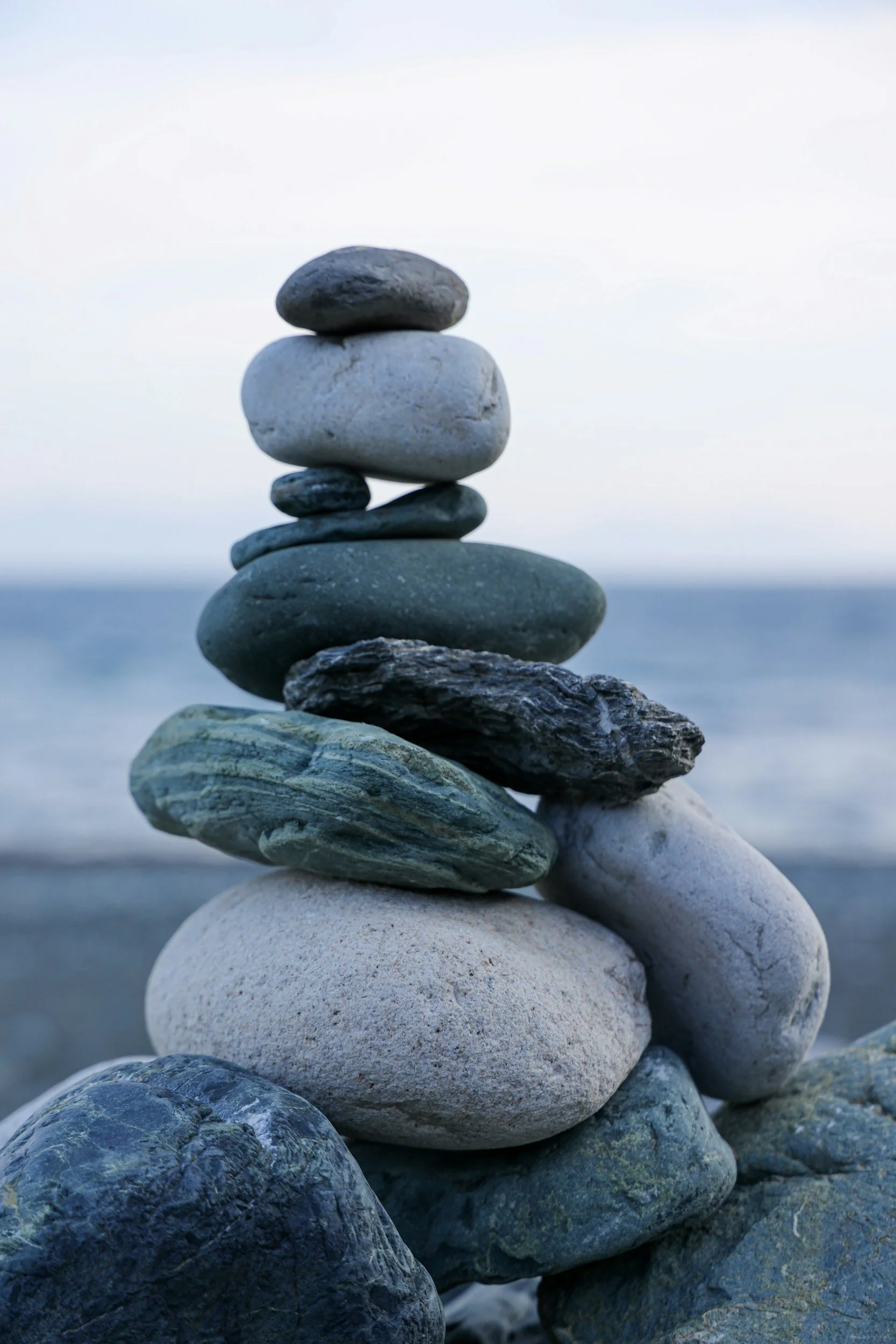 A vertical stack of various smooth, rounded stones of different sizes and colors, set against a blurry sky and ocean background.