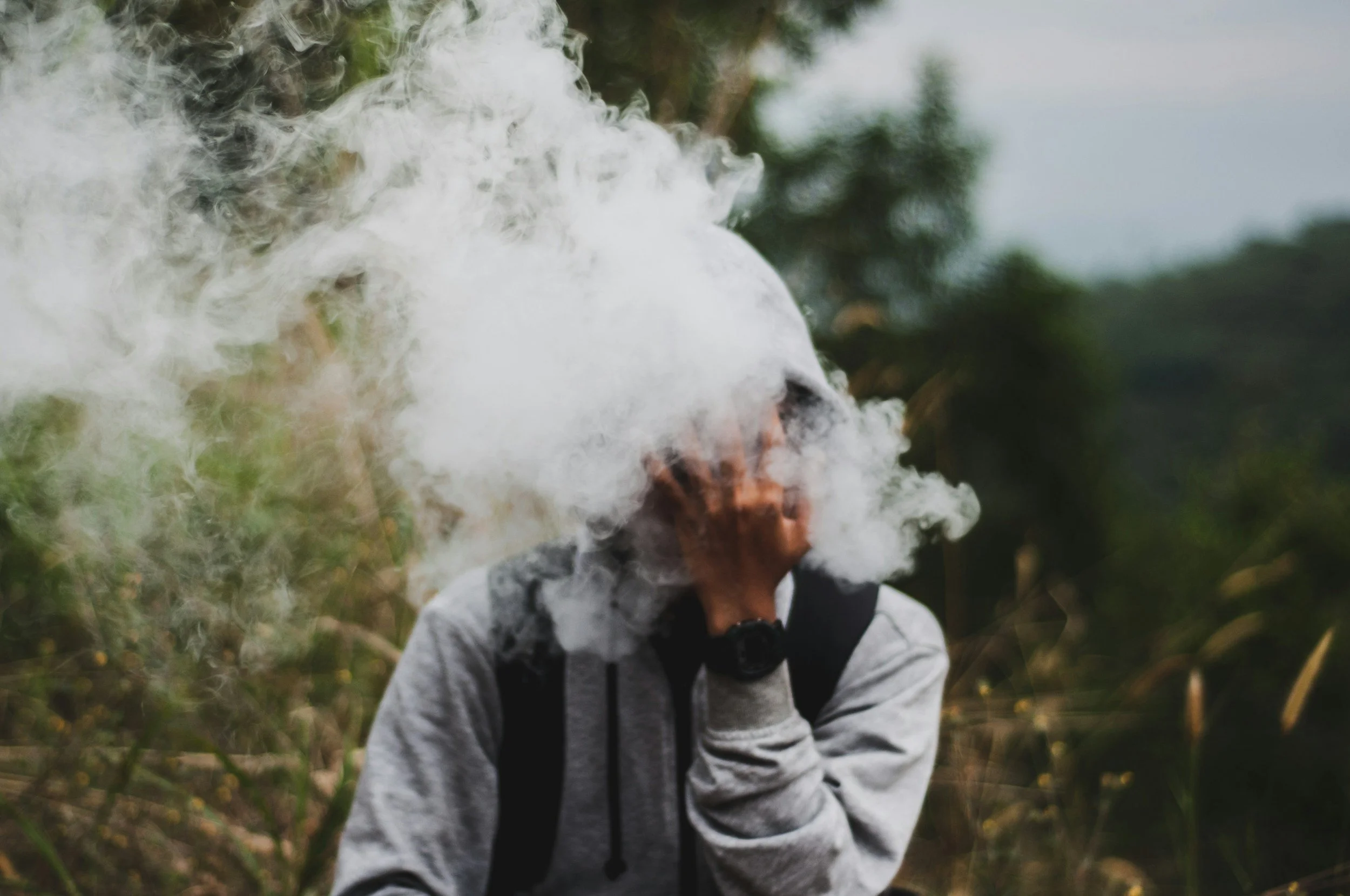 A person wearing a gray jacket and backpack exhaling white smoke outdoors with trees and greenery in the background.