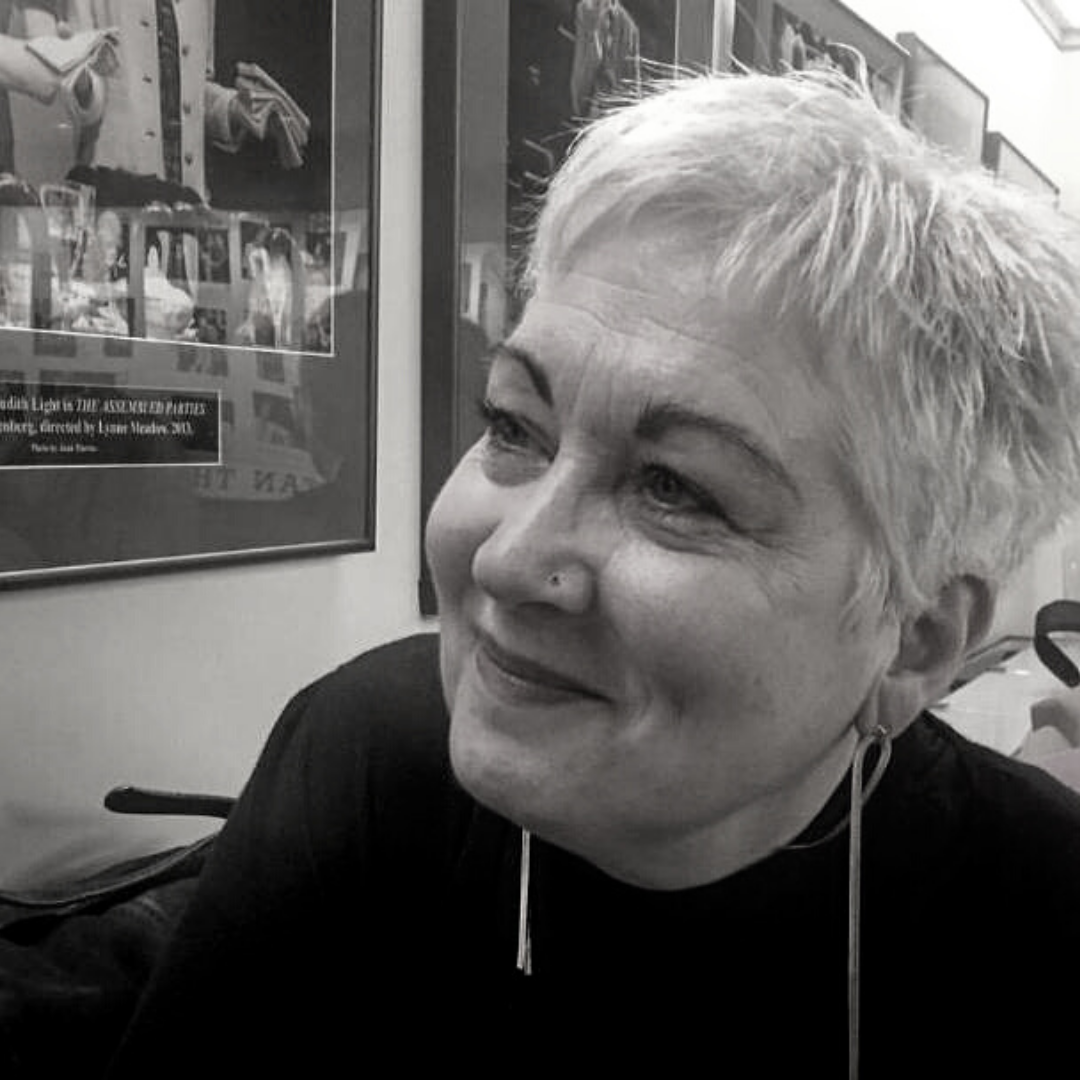 A woman with short, light-colored hair smiling and looking to the side, sitting indoors with framed pictures or posters on the wall behind her.