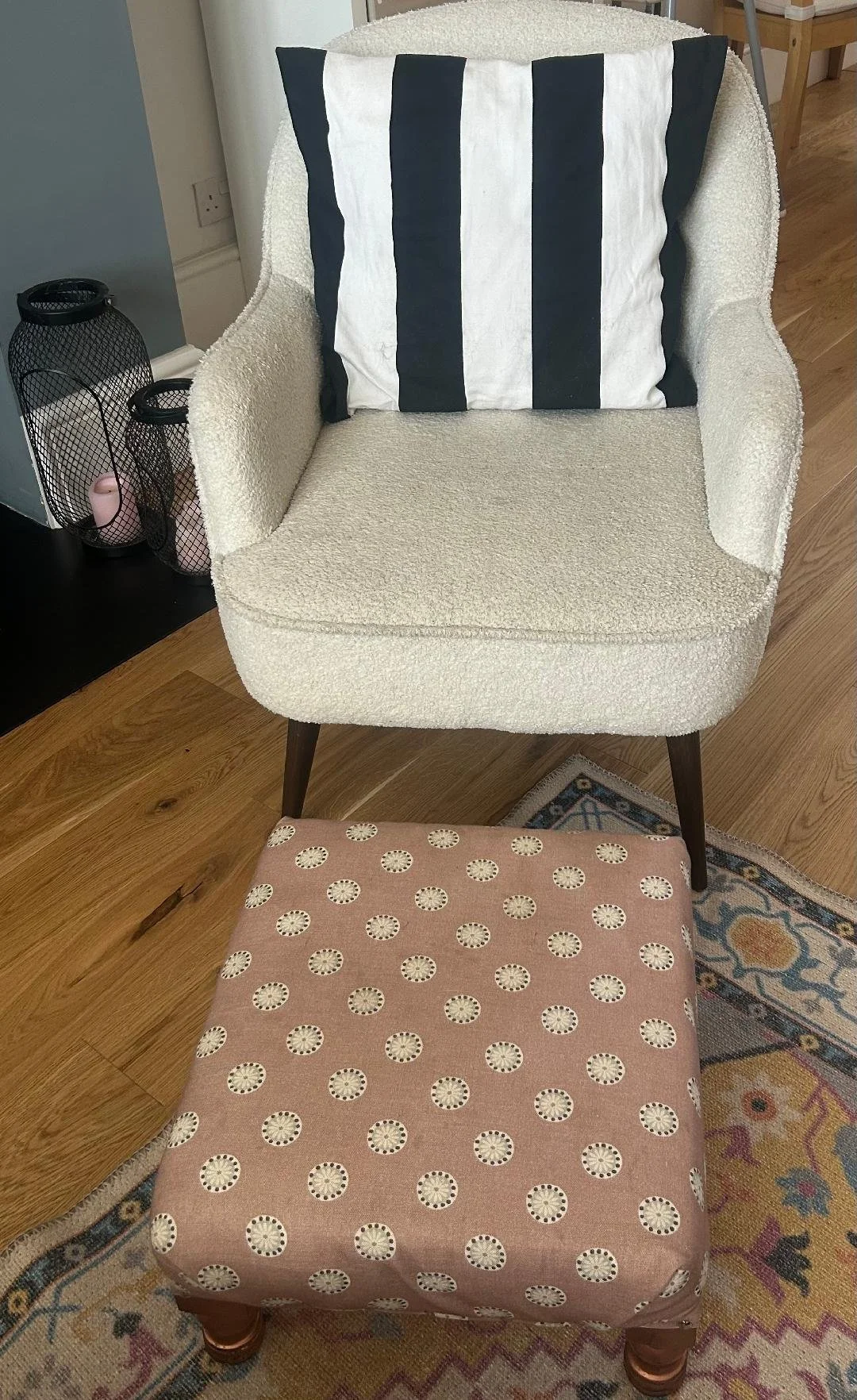 A beige upholstered armchair with dark wooden legs, paired with a pink footstool with a circular pattern, placed on a colorful patterned rug. A striped black and white pillow is on the armchair, which is near a small black wire basket with candles.